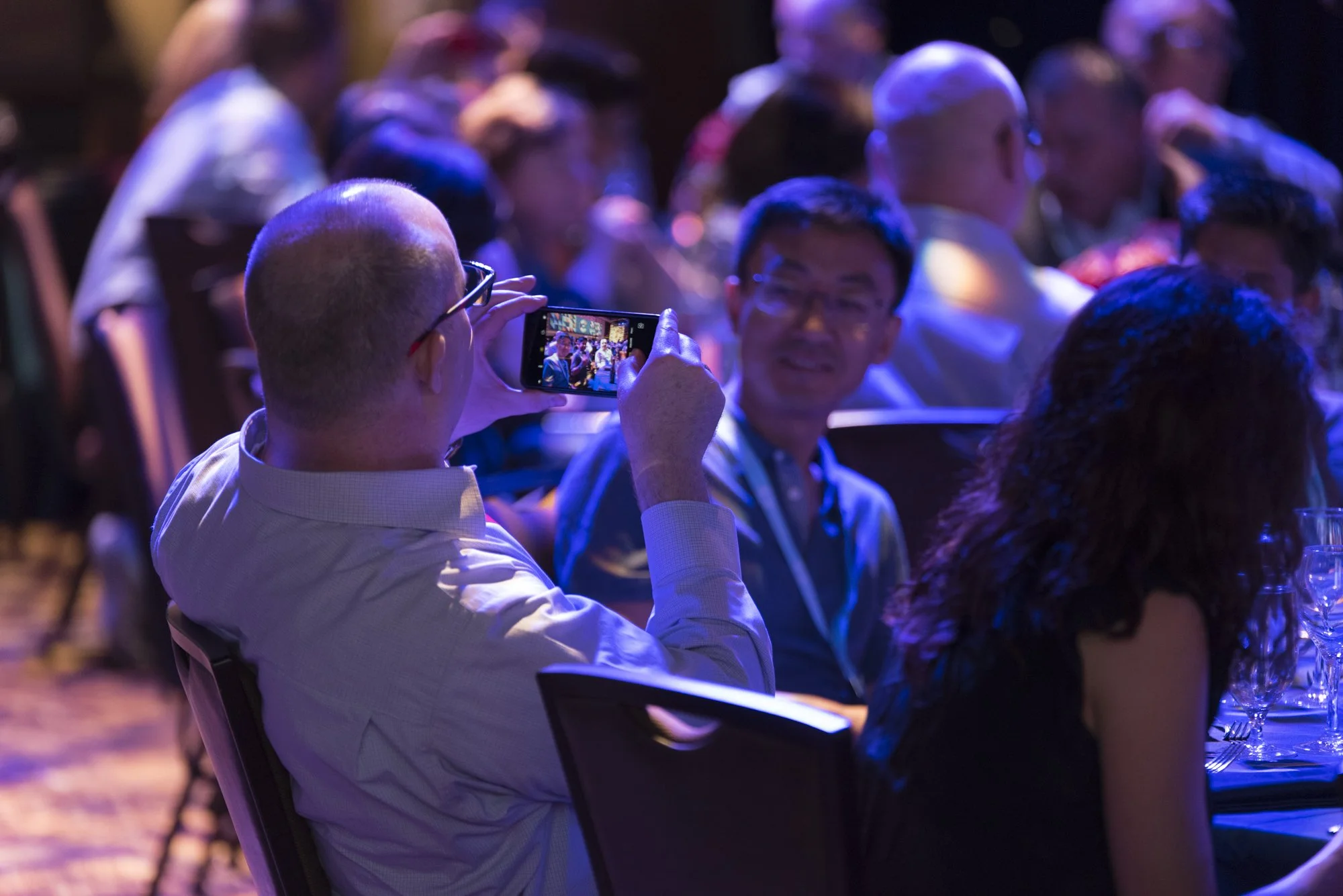 A man with glasses and a yellow shirt taking a photo with his smartphone at a crowded conference or event, with people seated at tables around him.