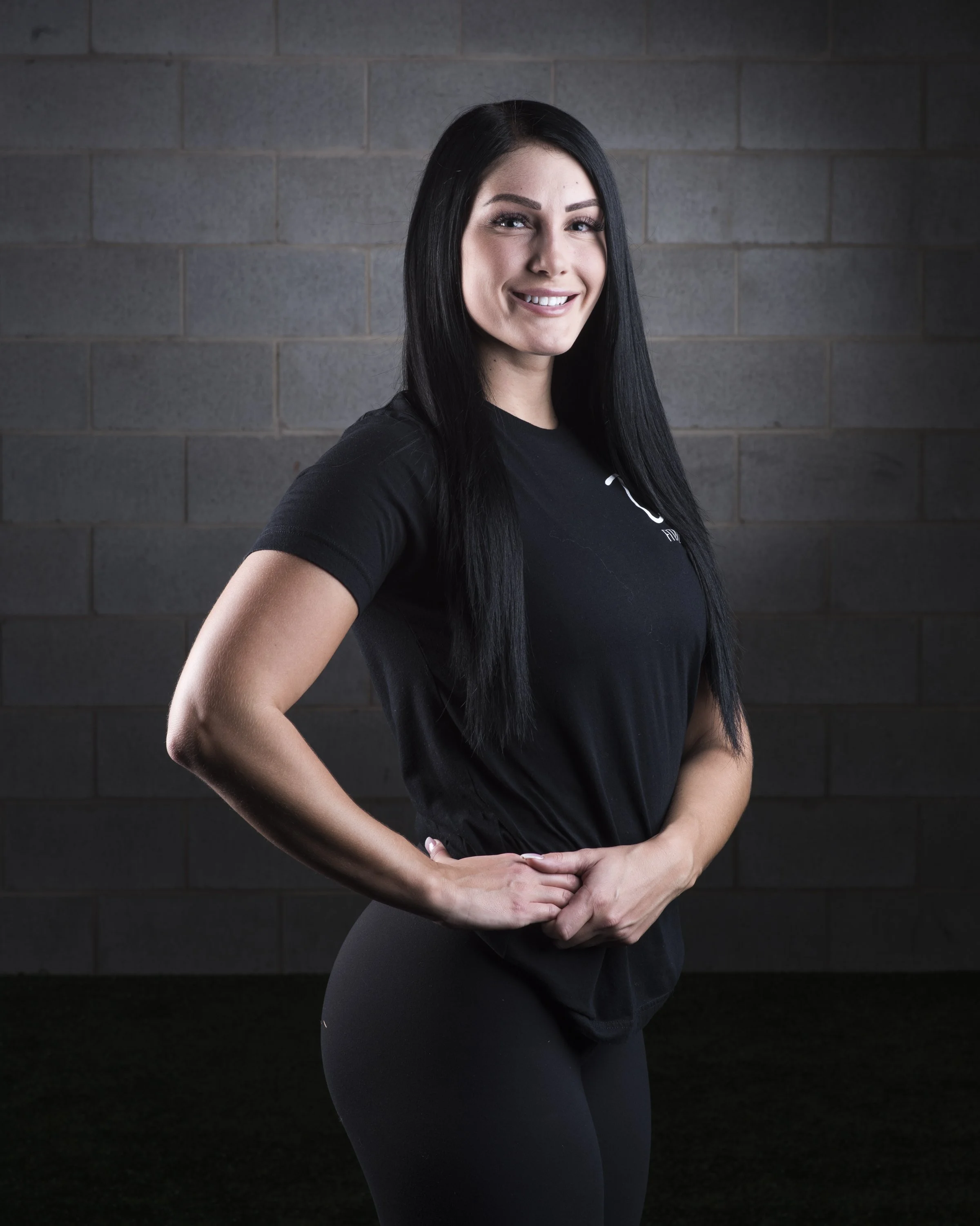 Woman with long black hair smiling, wearing black athletic clothing, standing against a gray brick wall.