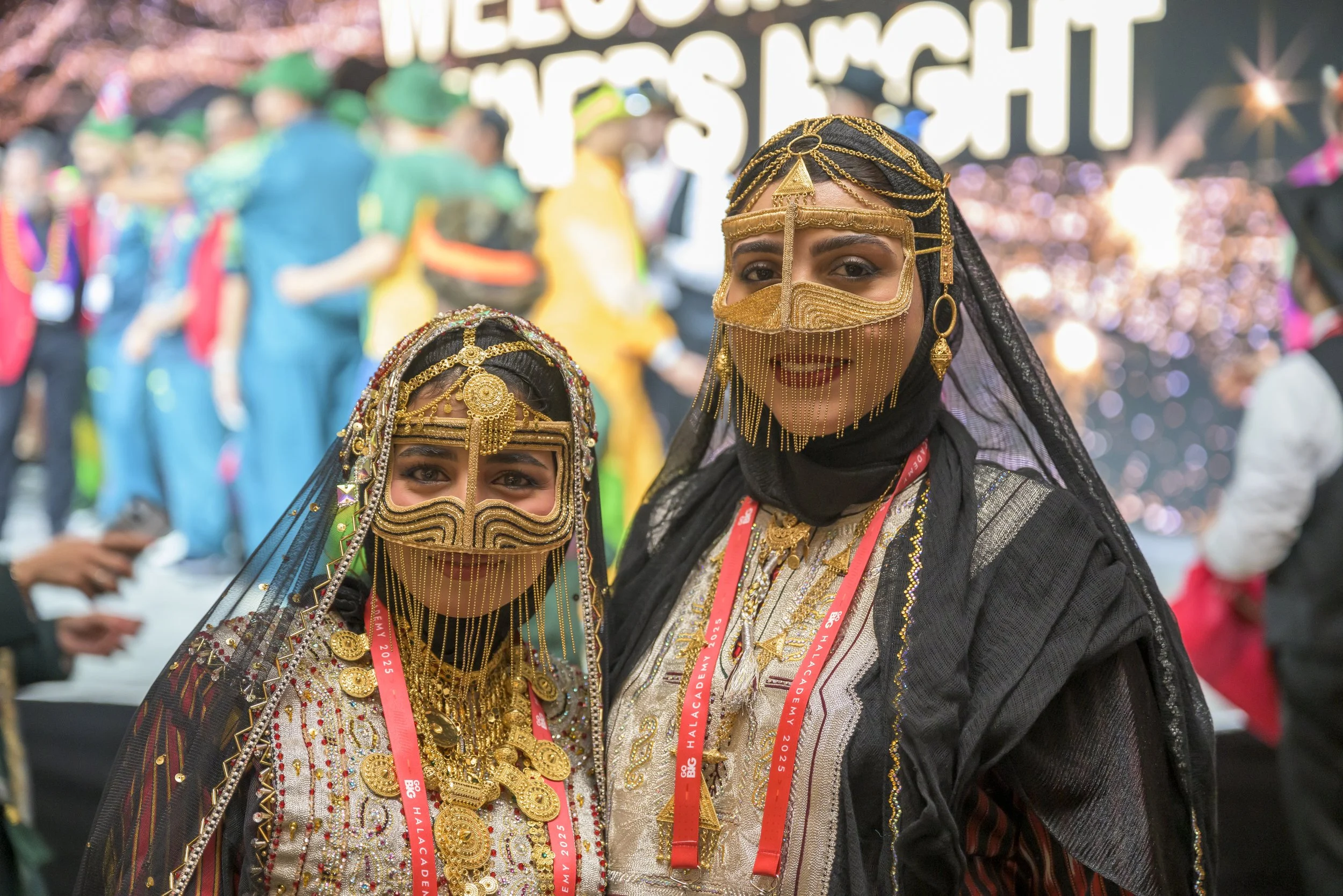 Two women wearing traditional African attire, gold jewelry, and ornate face masks at a cultural event.