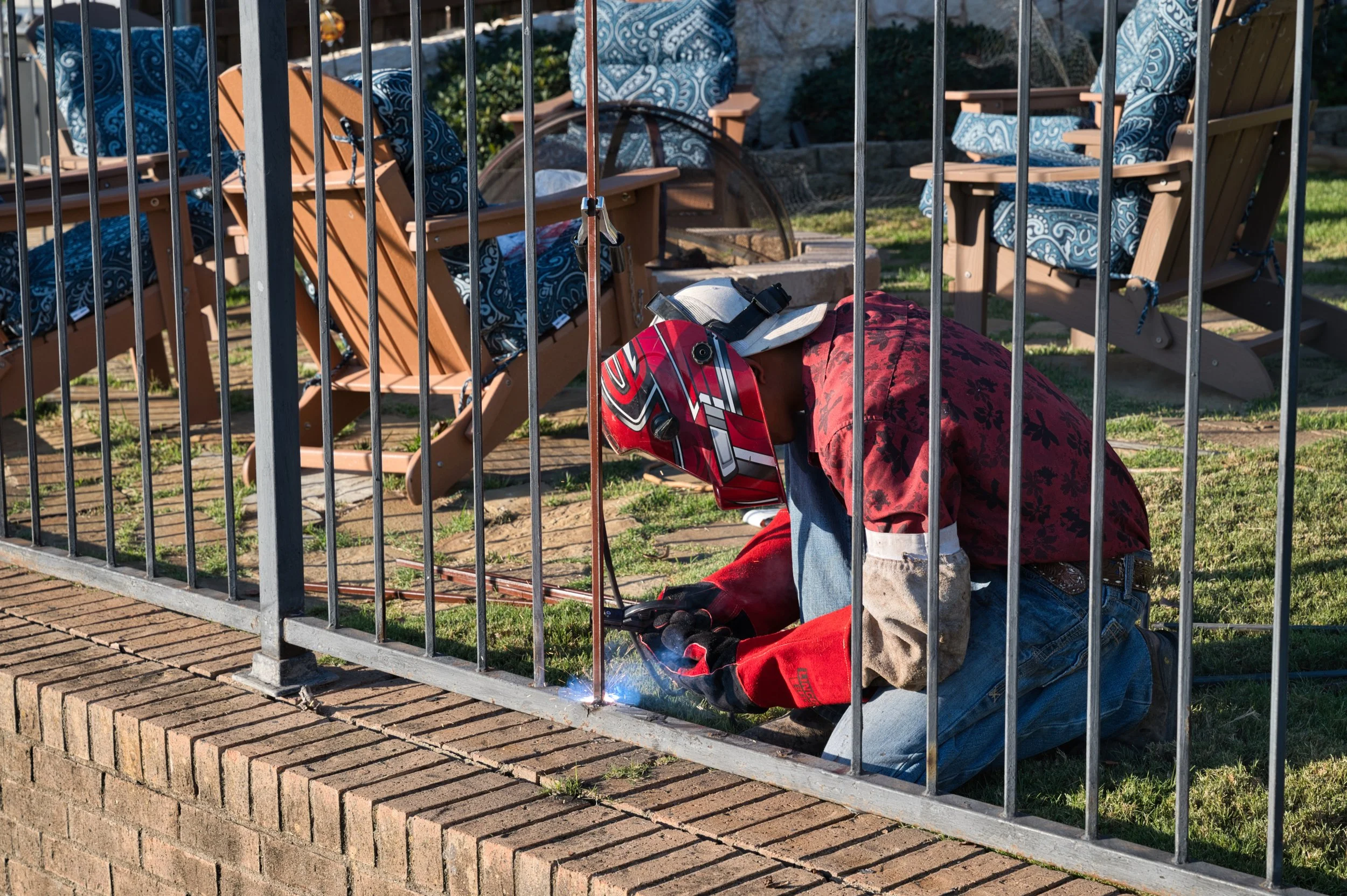 A person welding metal fence, wearing a red helmet and gloves, kneeling on the ground next to a brick wall with wooden chairs and tables in the background.