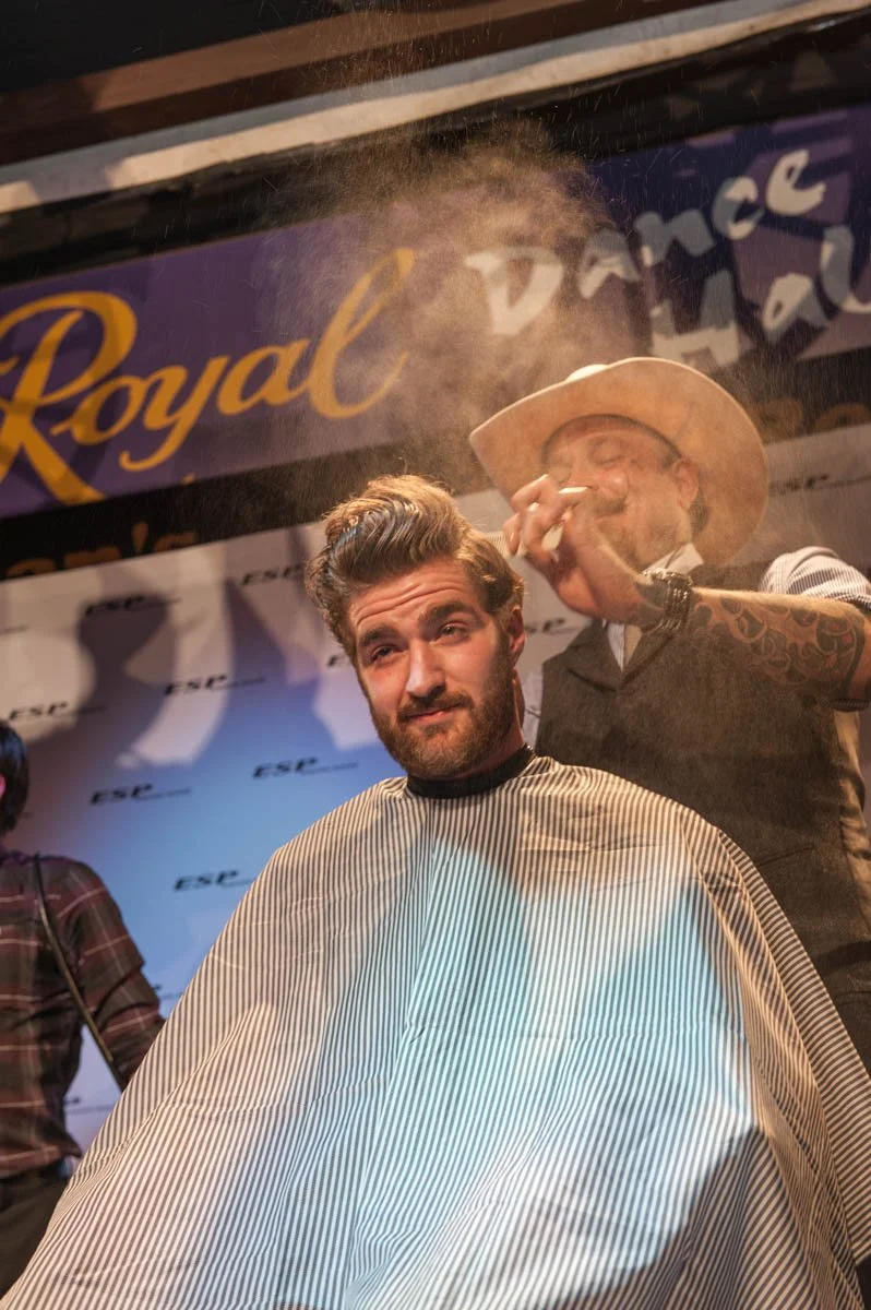 A man getting a haircut in a barber shop with a man in a cowboy hat smoking a cigar in the background.