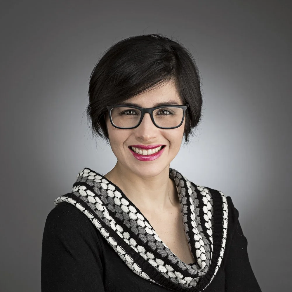 A woman with short dark hair, wearing glasses, a black top, and a patterned scarf, smiling against a gray background.