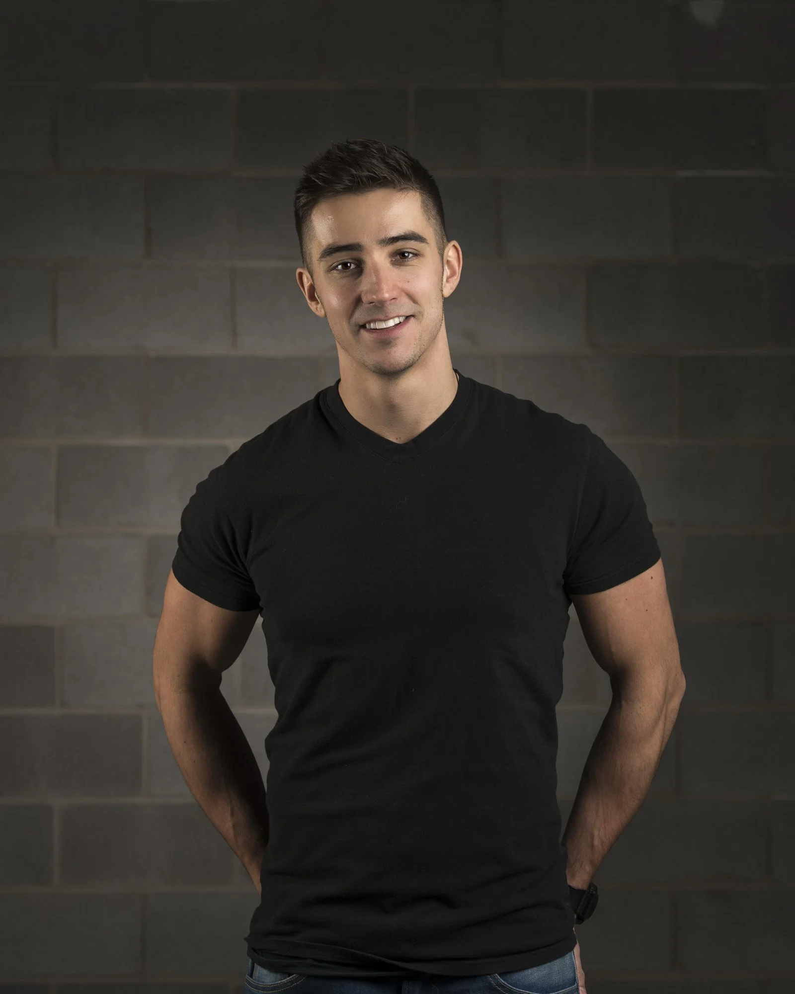 A young man with short dark hair smiling, wearing a black t-shirt and blue jeans, standing against a dark gray brick wall background.