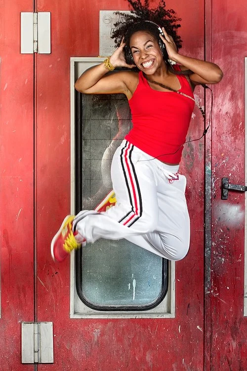 A woman wearing a red tank top, white sweatpants with red and black stripes, and yellow sneakers is jumping in front of a red door while listening to music with headphones.