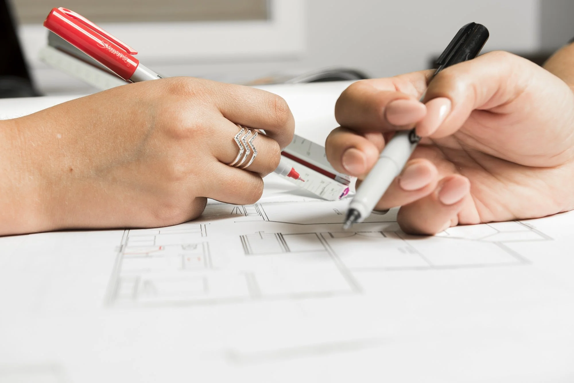 Two people working on an architectural blueprint, one holding a red pen and the other holding a black pen, with a measuring tool nearby.