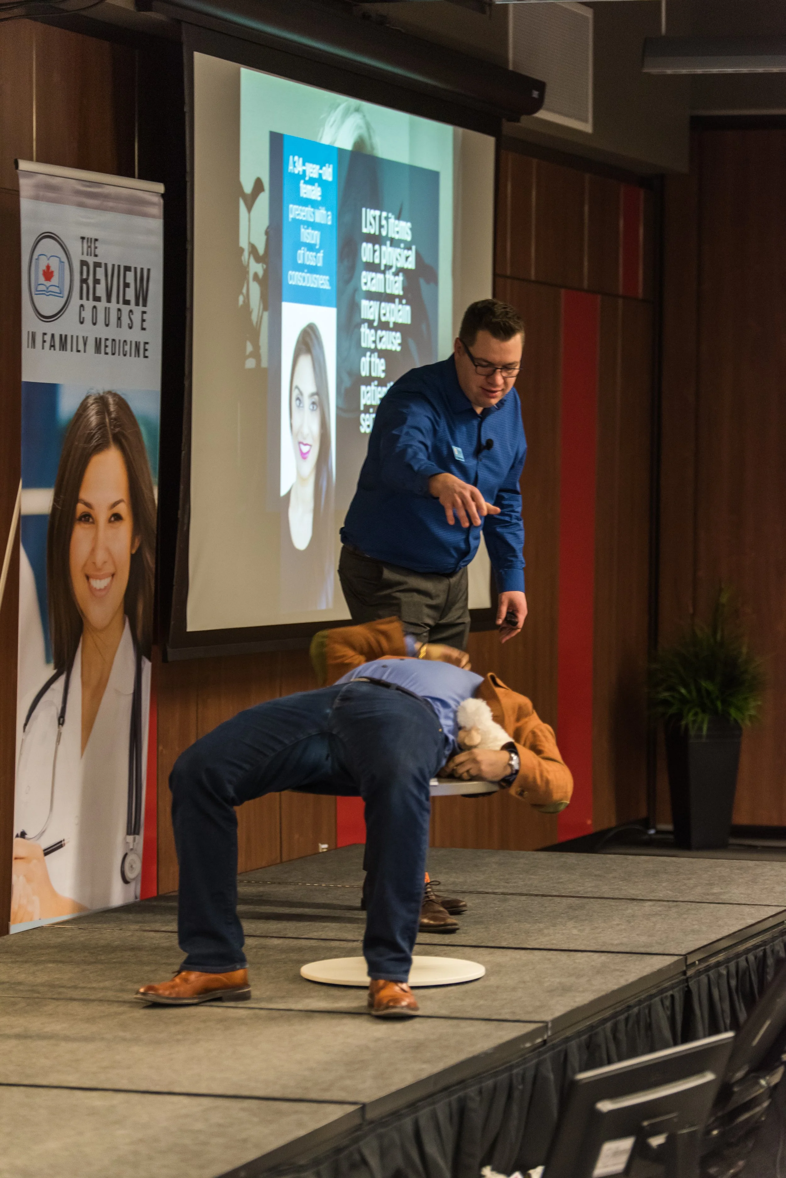 A presenter on stage demonstrating a medical procedure with a man bent over holding a medical plush toy and a chatty man in a blue shirt pointing at him.