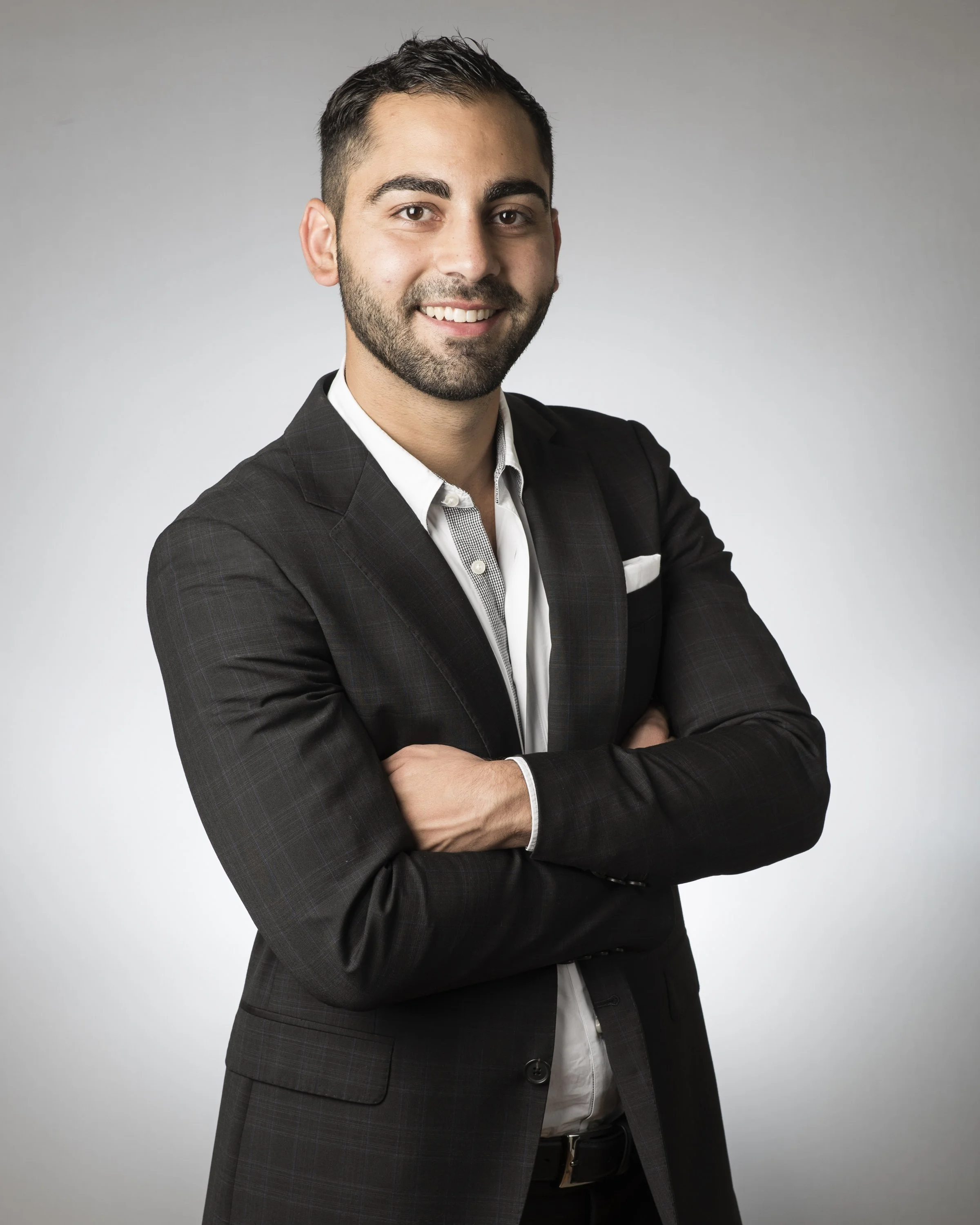 A man in a dark checkered suit with a white shirt, crossed arms, smiling, posing against a light gray background.