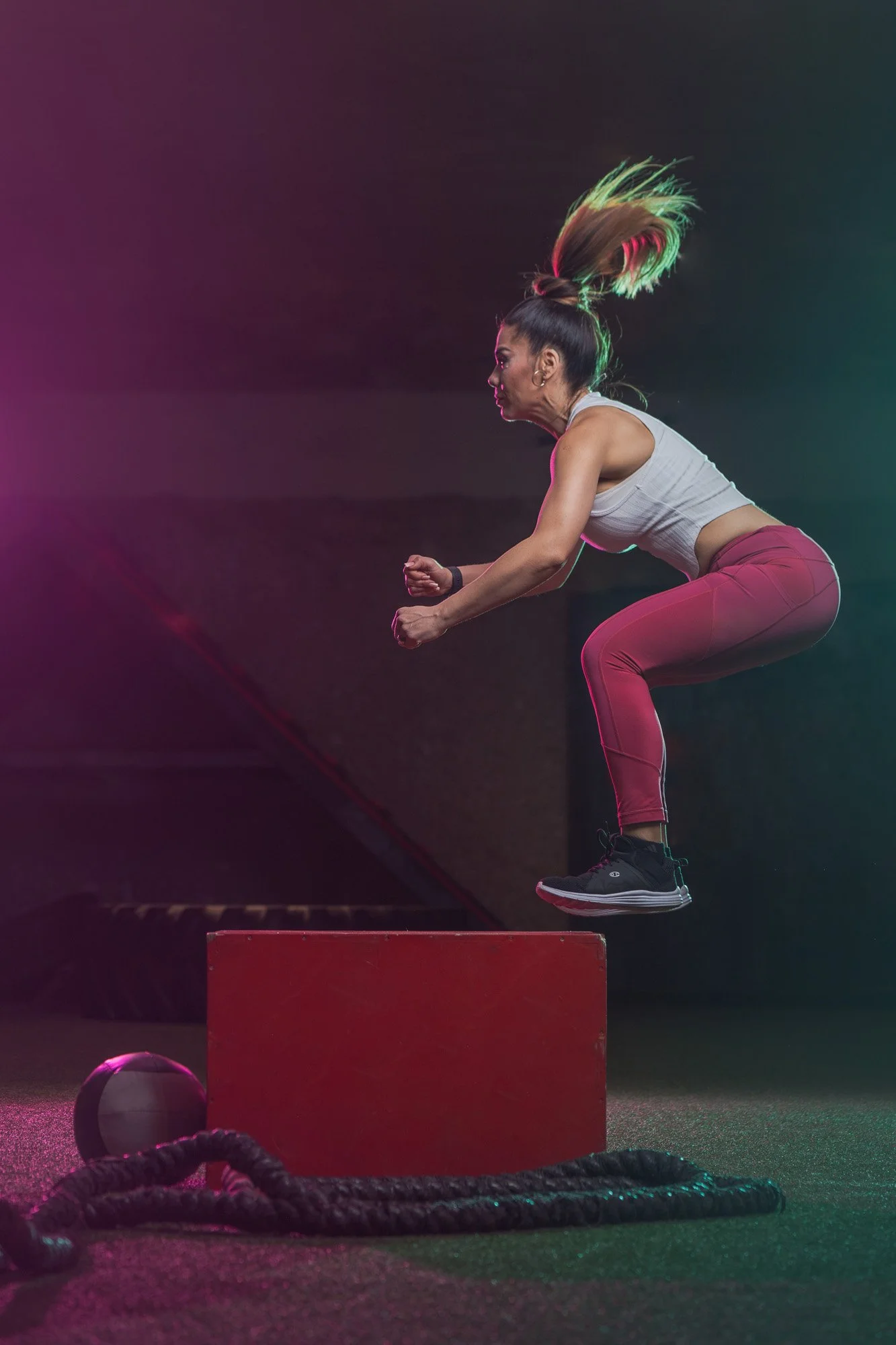 A woman with blonde hair tied in a high ponytail appears to be jumping onto a red block in a gym. She is wearing a white sports bra and pink leggings, with fitness equipment such as a medicine ball and battle ropes on the ground nearby.