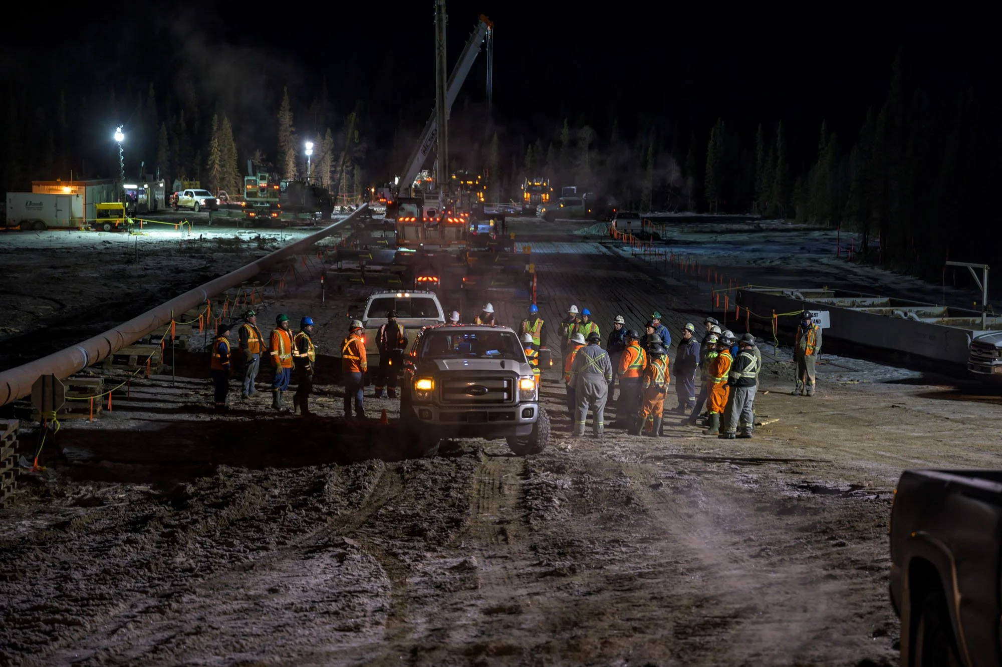 Nighttime construction site with numerous workers in safety vests and helmets gathered near a parked truck, multiple construction vehicles, and equipment, with a crane lifting a large pipe, and several lights illuminating the area.