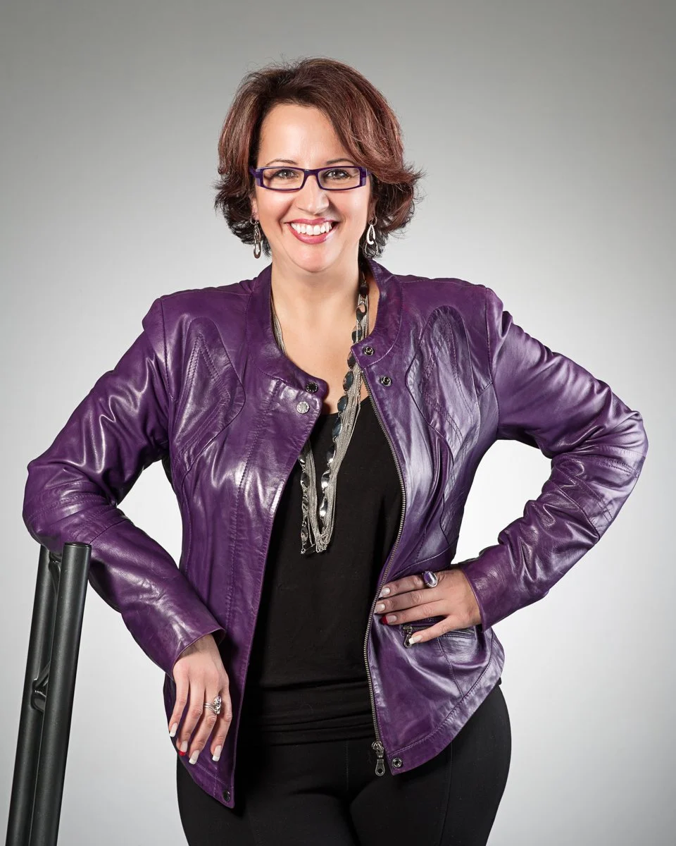 Woman with short brown hair and glasses in a purple leather jacket, smiling, against a plain gray backdrop.
