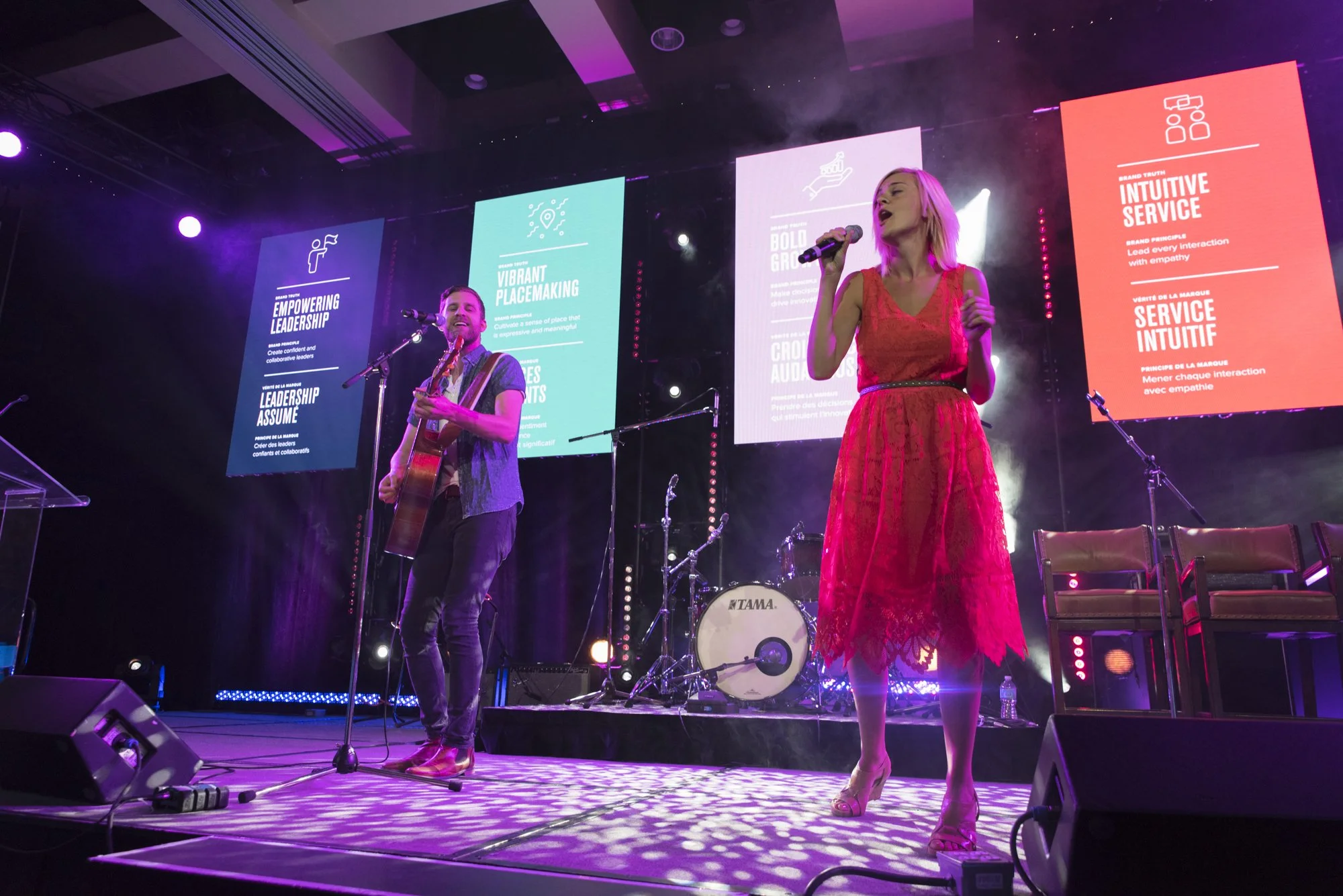 A man playing guitar and a woman singing on stage during a concert, with colorful presentation slides projected behind them.