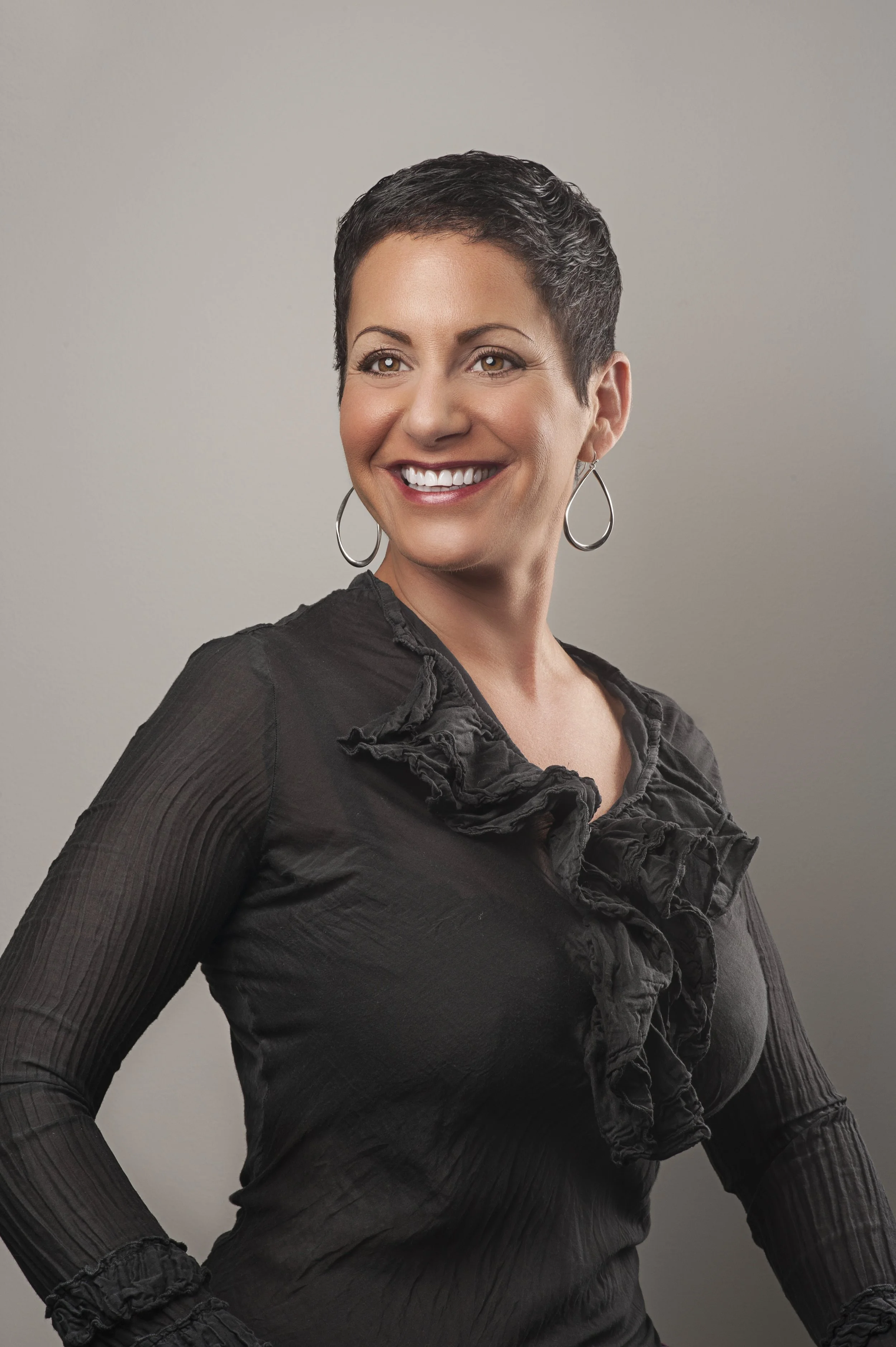 A woman with short dark hair, wearing a black blouse with ruffled details and large hoop earrings, smiling and posing against a plain background.