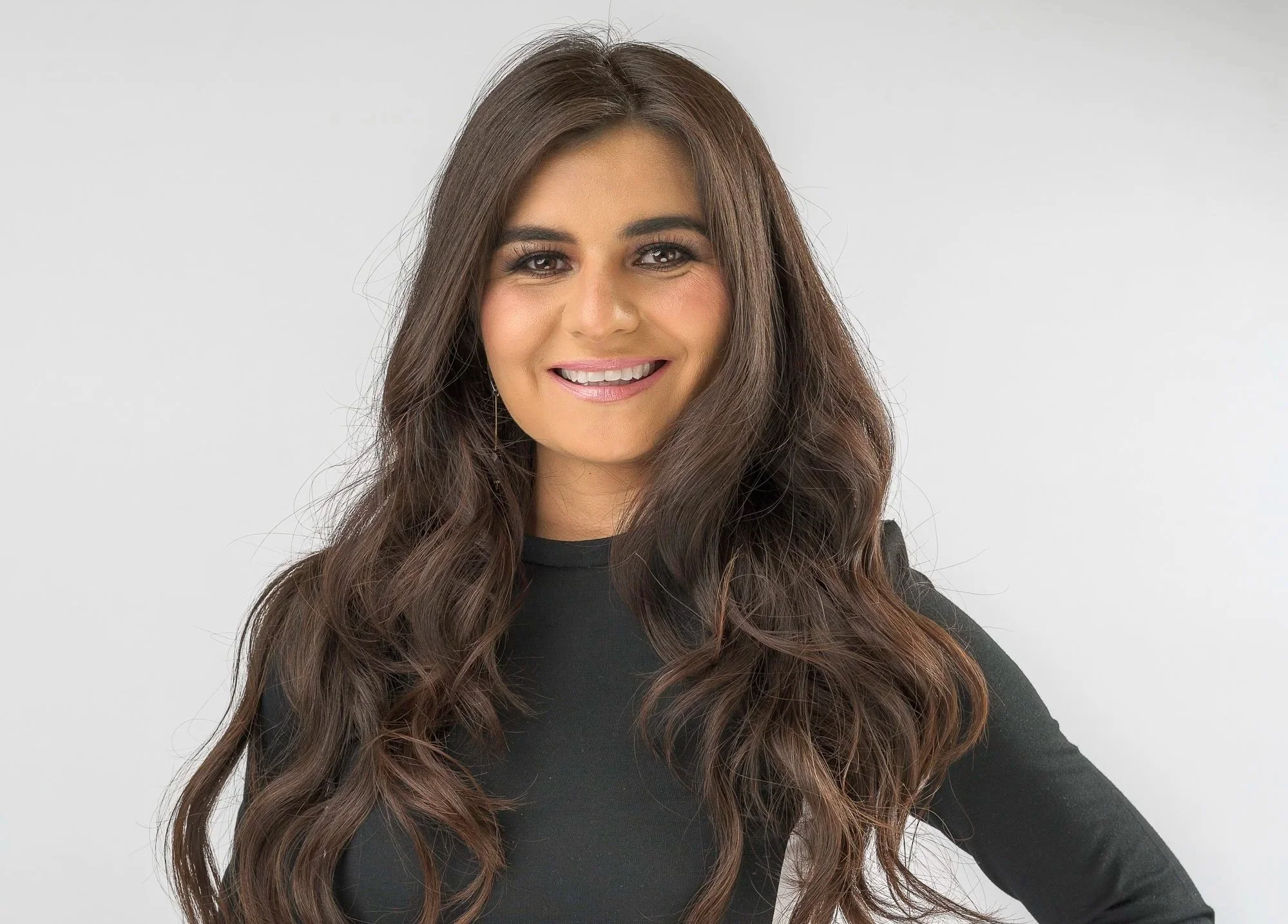A young woman with long, wavy brown hair smiling, wearing a black top, standing against a plain white background.