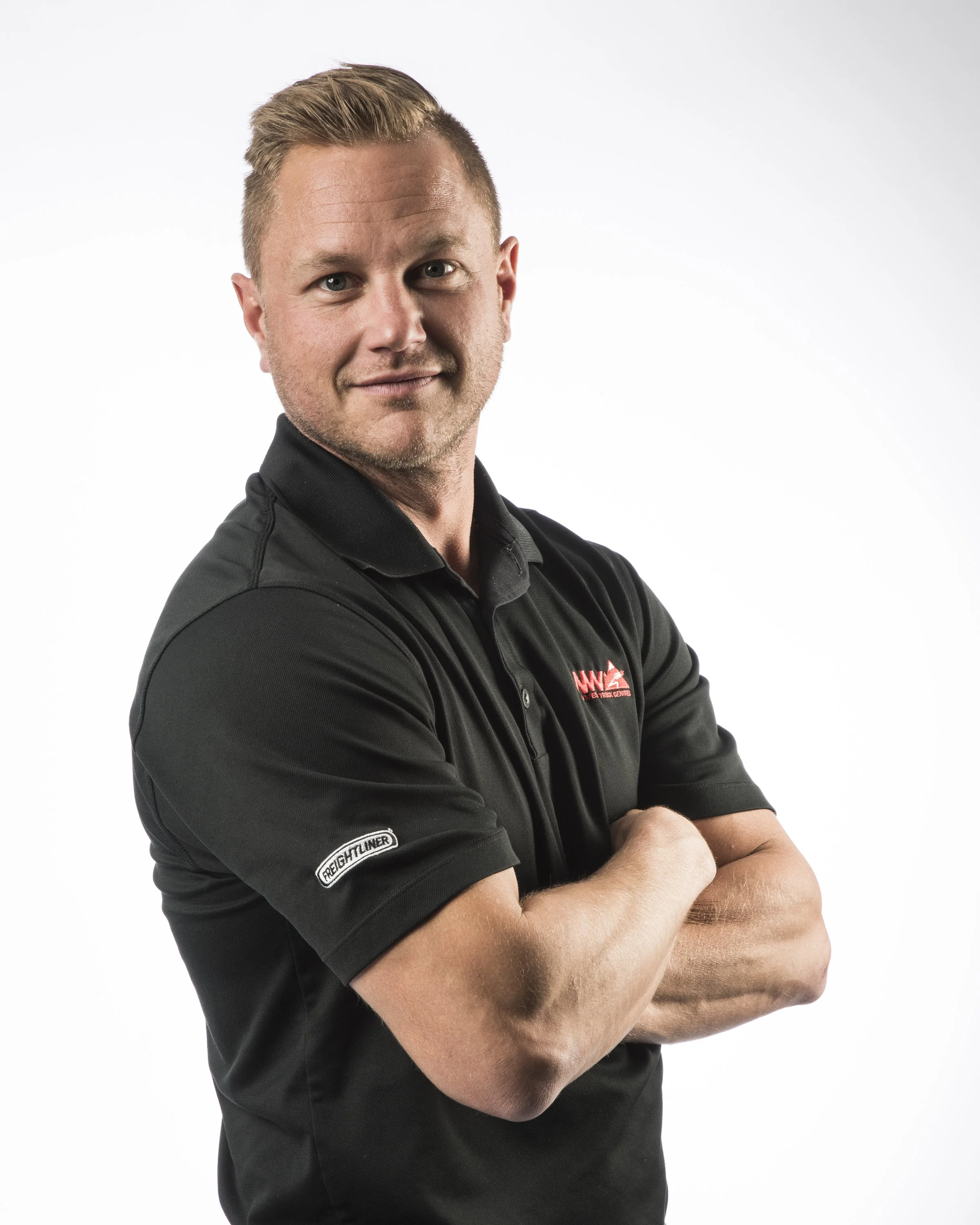 A man with short blond hair wearing a black polo shirt with logos, standing with arms crossed against a plain white background.