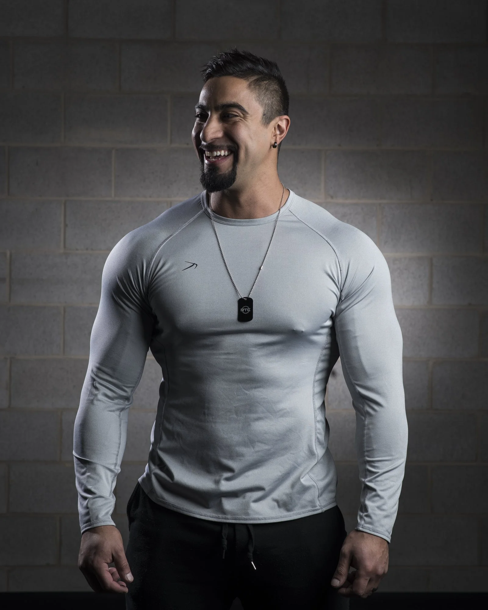 A smiling man with dark hair, a beard, and an earring, wearing a tight-fitting light gray athletic shirt and a black dog tag necklace, standing against a gray brick wall.