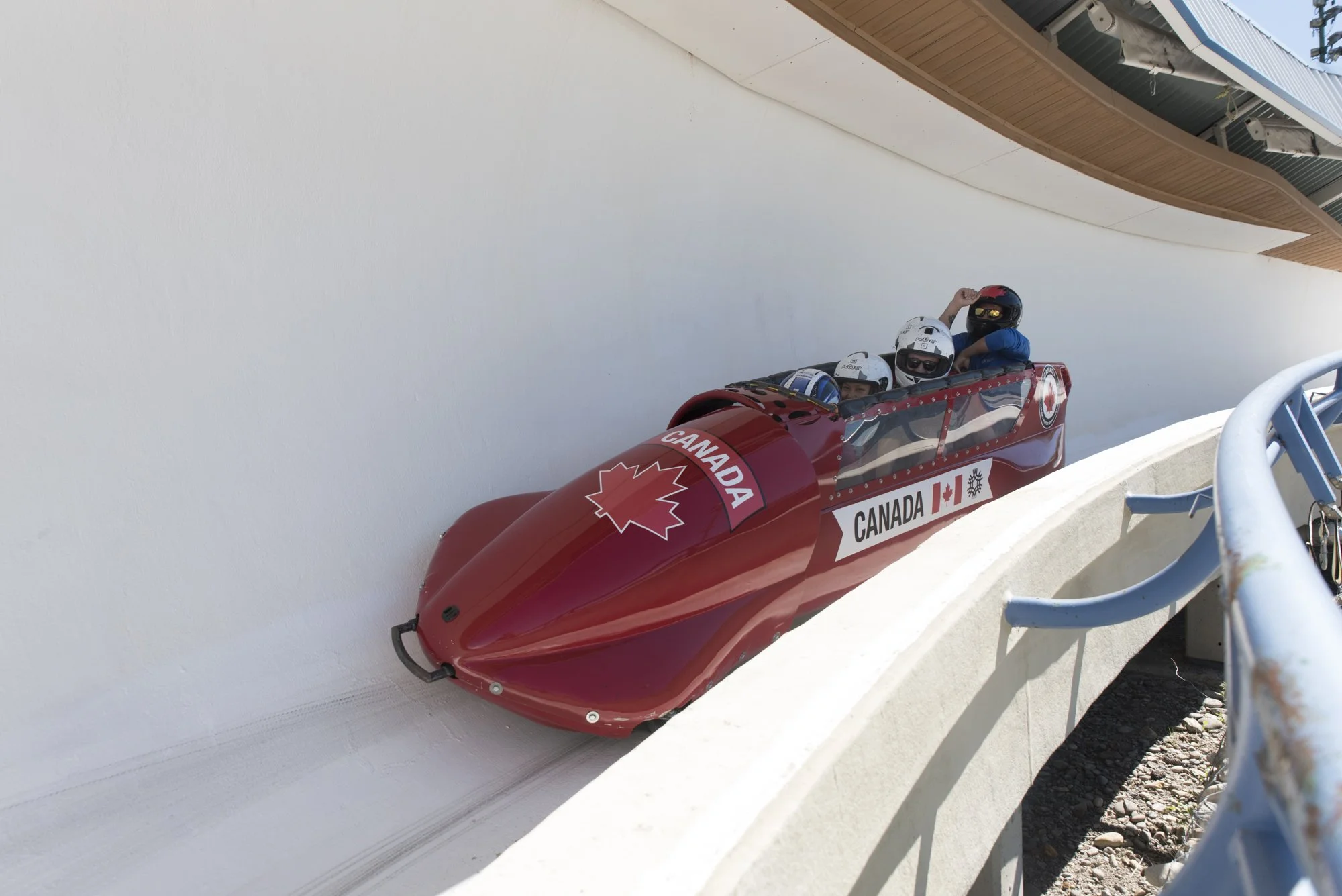 A red Canadian bobsled with four people inside, wearing helmets, navigating a twisty bobsled track.