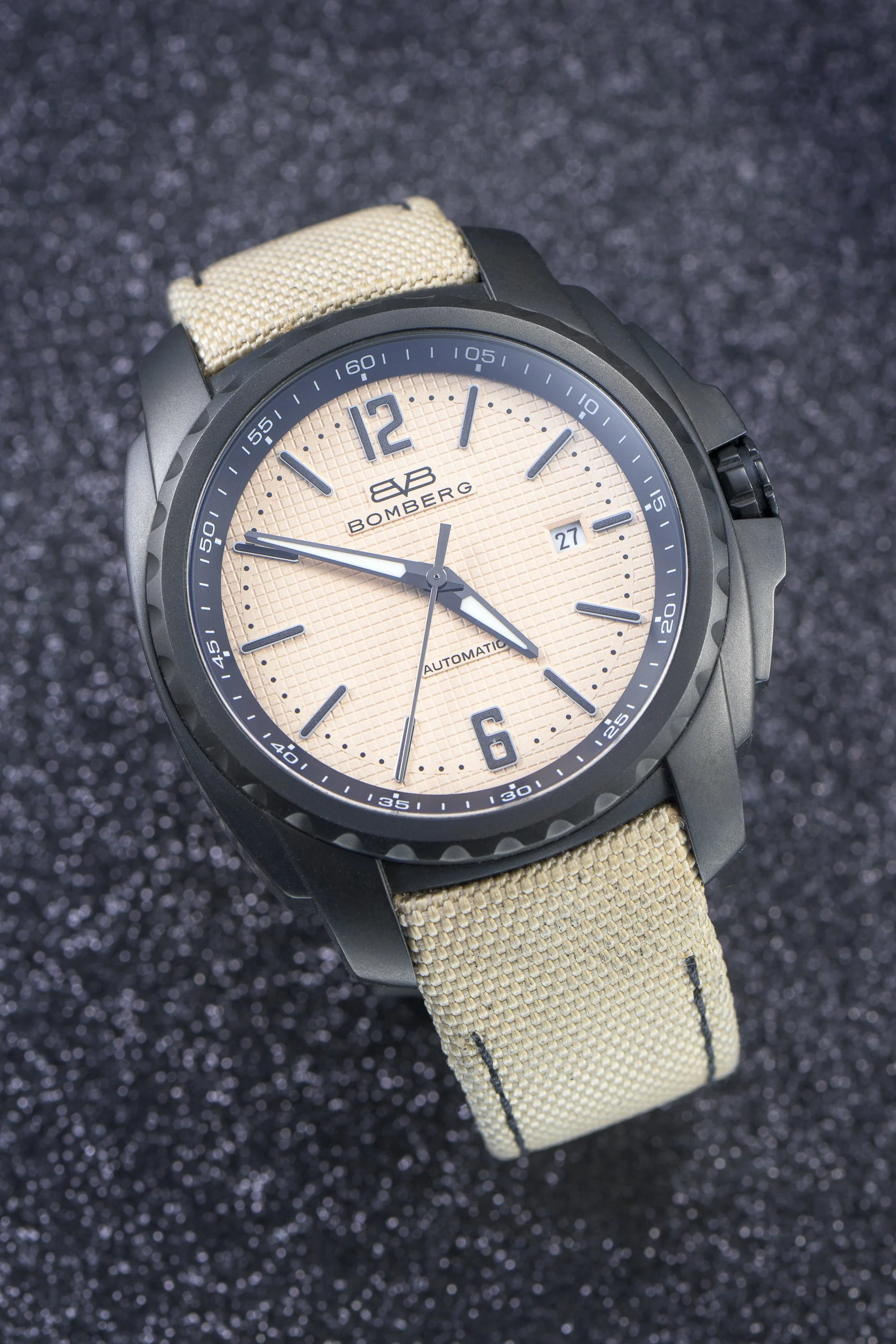 A close-up view of a wristwatch with a black case, beige textured dial, and a beige fabric strap. The watch displays the time with silver hour, minute, and second hands, and has a date window at 3 o'clock. The dial features the branding 'Bombberg' an