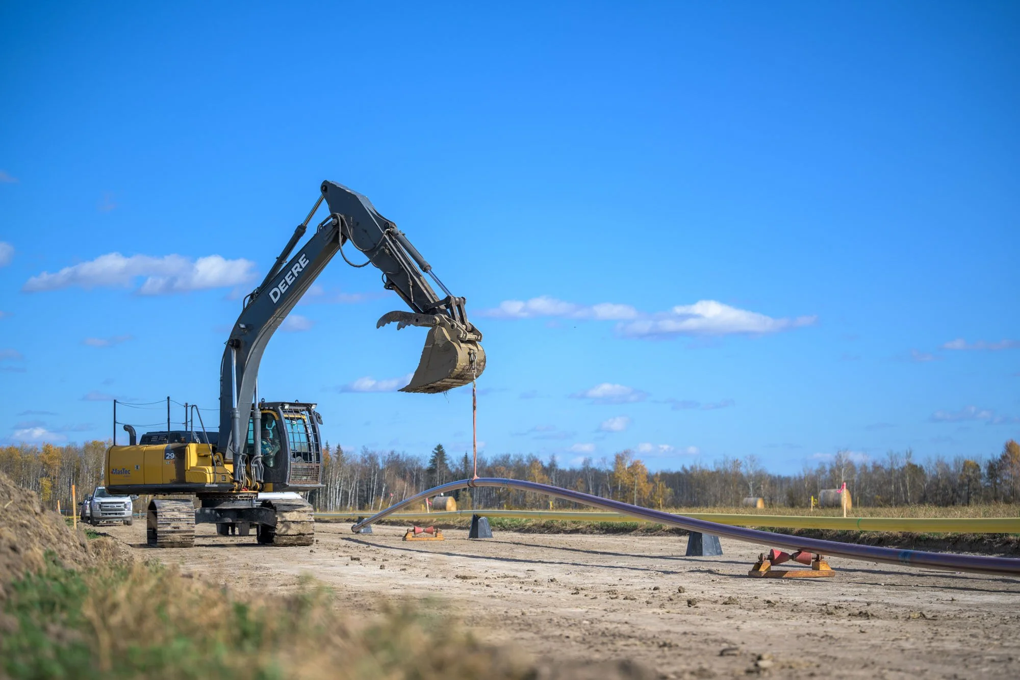 A construction excavator lifting a pipe at a construction site on a clear day with a blue sky and scattered clouds.