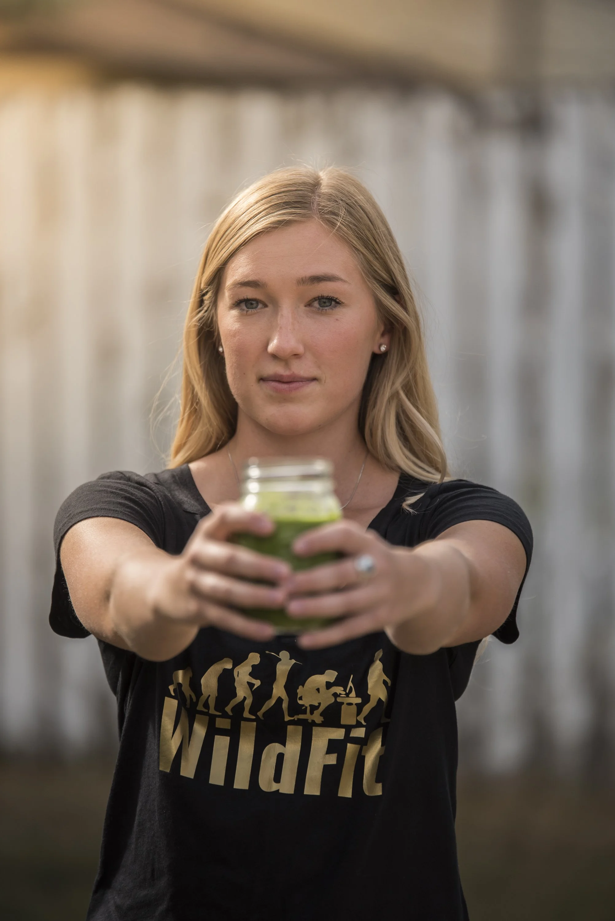 A woman with blonde hair and a black T-shirt holding a small glass jar of green smoothie or juice, extending it forward.