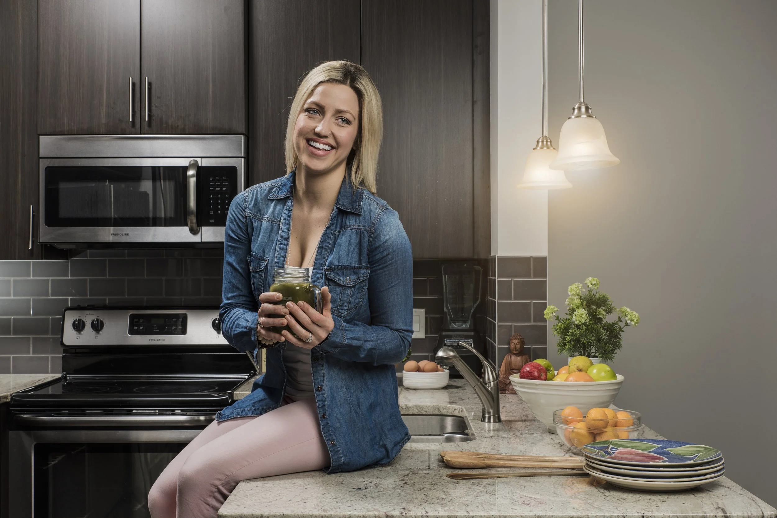 A woman sitting on a kitchen counter holding a jar of green smoothie, smiling, with a modern kitchen background including a microwave, stove, and bowls of fruit.