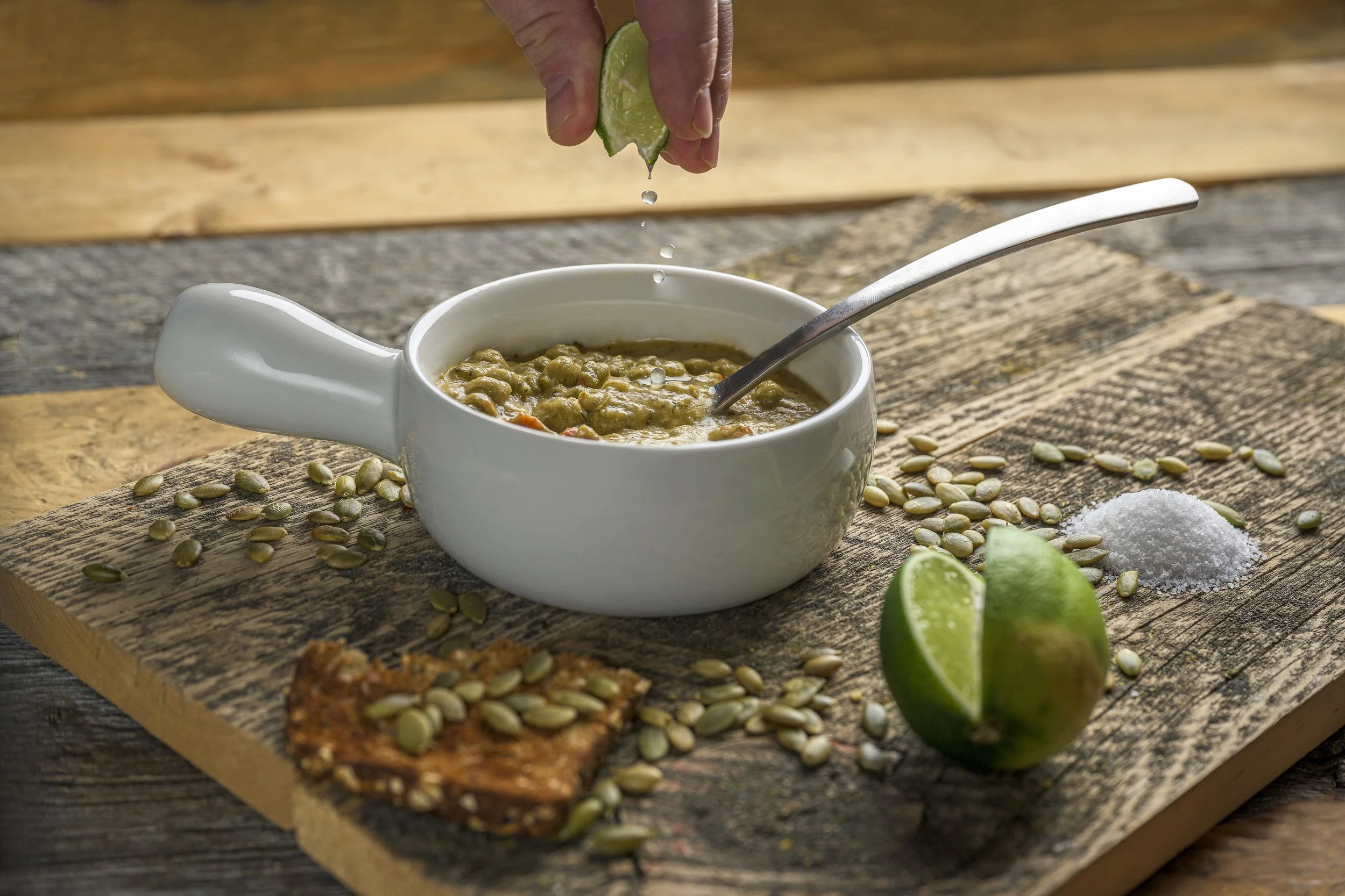 A hand squeezing a lime over a bowl of soup on a rustic wooden surface. Pumpkin seeds, a salt pile, a lime wedge, and a fried tortilla chip are also on the surface.