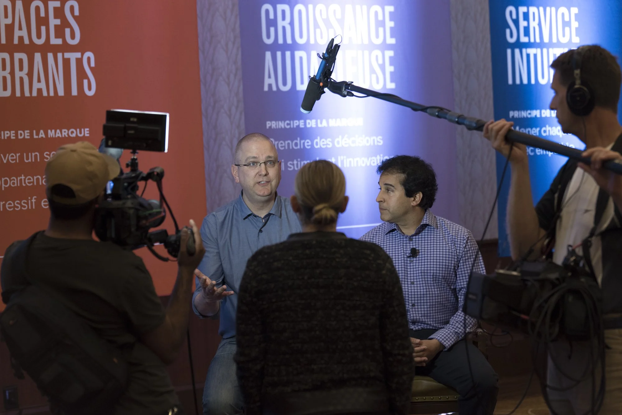 Two men and two women seated during an interview or discussion, surrounded by cameras and a boom microphone in a room with colorful posters in the background.