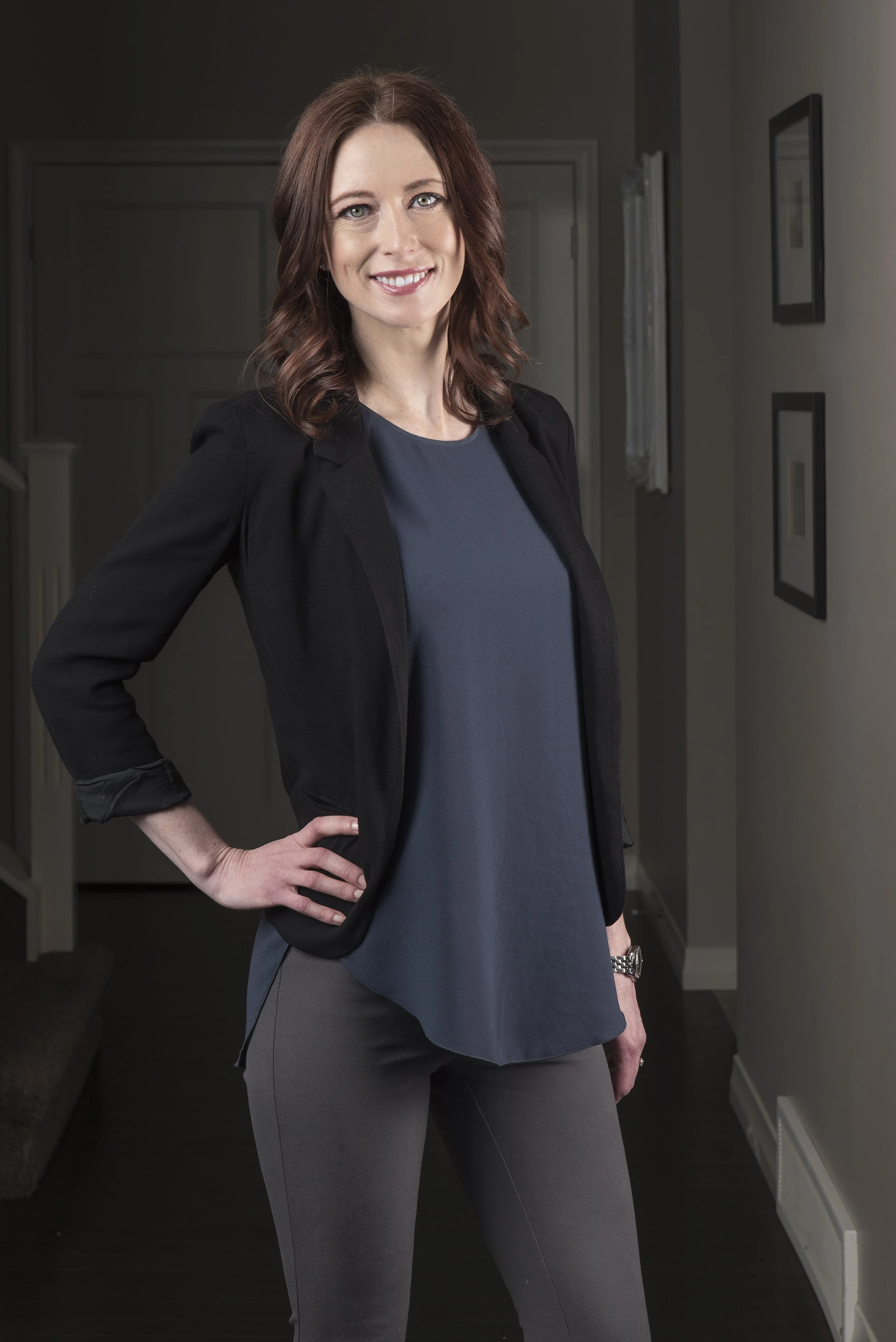 A woman with wavy, shoulder-length brown hair standing indoors with her right hand on her hip. She is wearing a black blazer, a navy blue top, and gray pants. She is smiling and looking at the camera in front of a wall decorated with framed pictures.