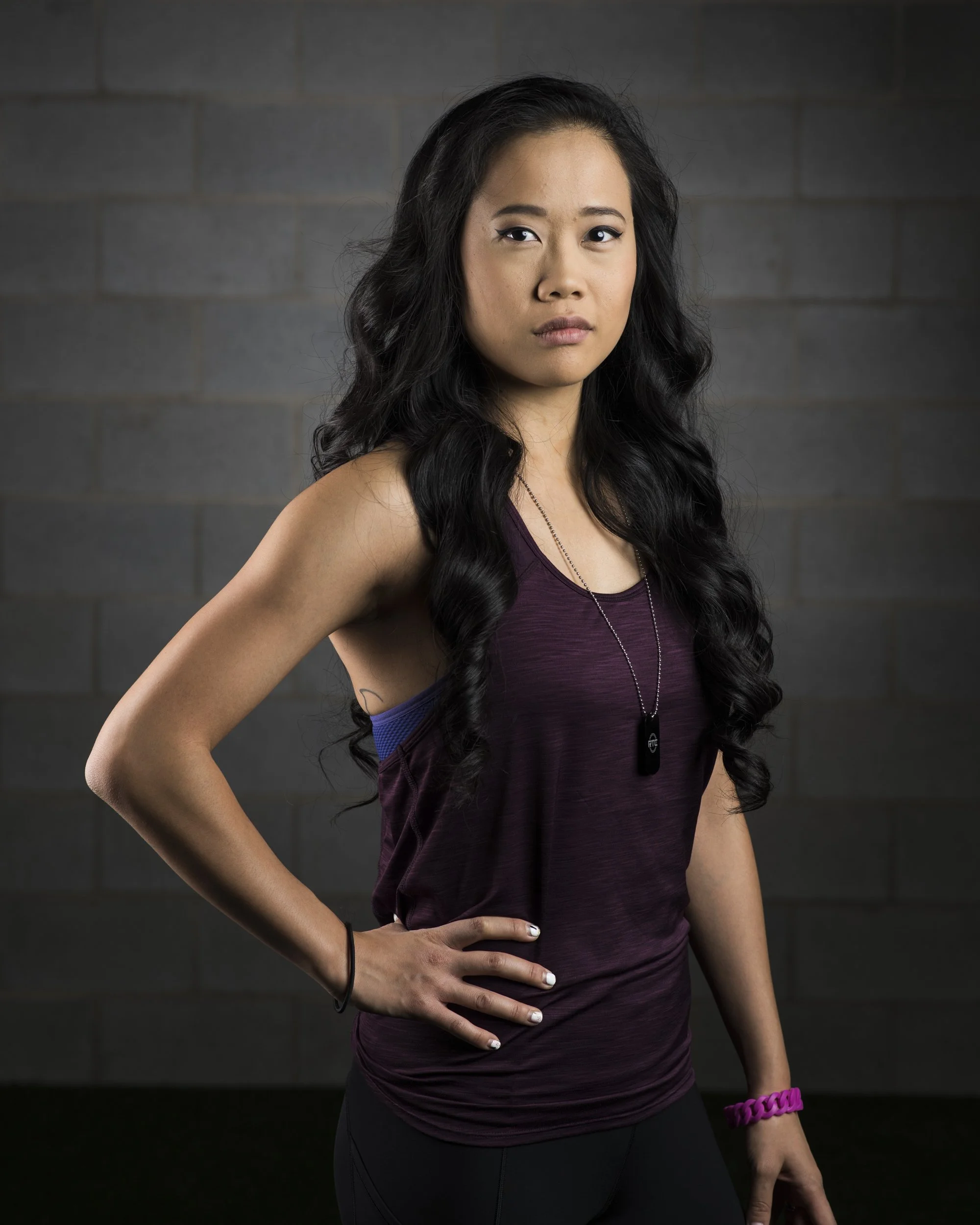 A young woman with long black wavy hair wearing a purple tank top, black leggings, a necklace with a black dog tag, a pink bracelet, and a black hair tie, standing with one hand on her hip against a gray brick wall.