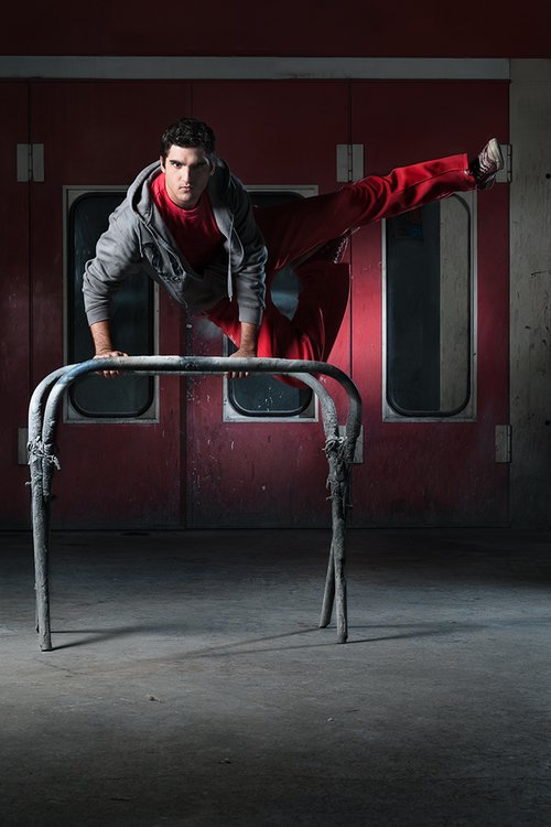 A young man performing a parkour move in an indoor gym, balancing on a metal hurdle with one hand while extending one leg out to the side.