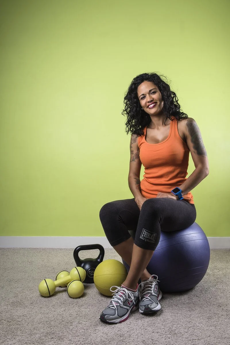 A woman sitting on a blue exercise ball in a home gym with green walls, wearing a sleeveless orange top, black leggings, and gray athletic shoes. She is smiling and has curly black hair. There are workout items on the floor, including a yellow medici