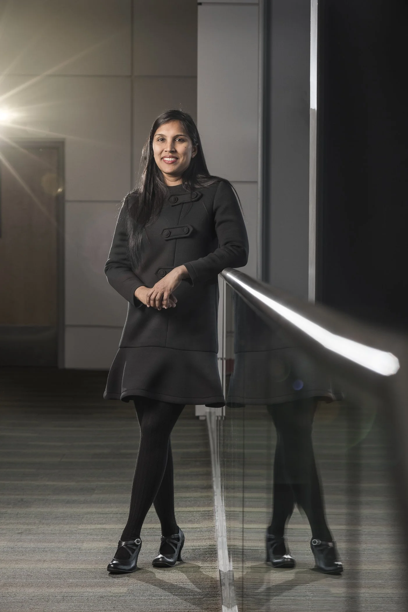 A professional woman in a black outfit standing indoors near a glass railing, smiling at the camera.