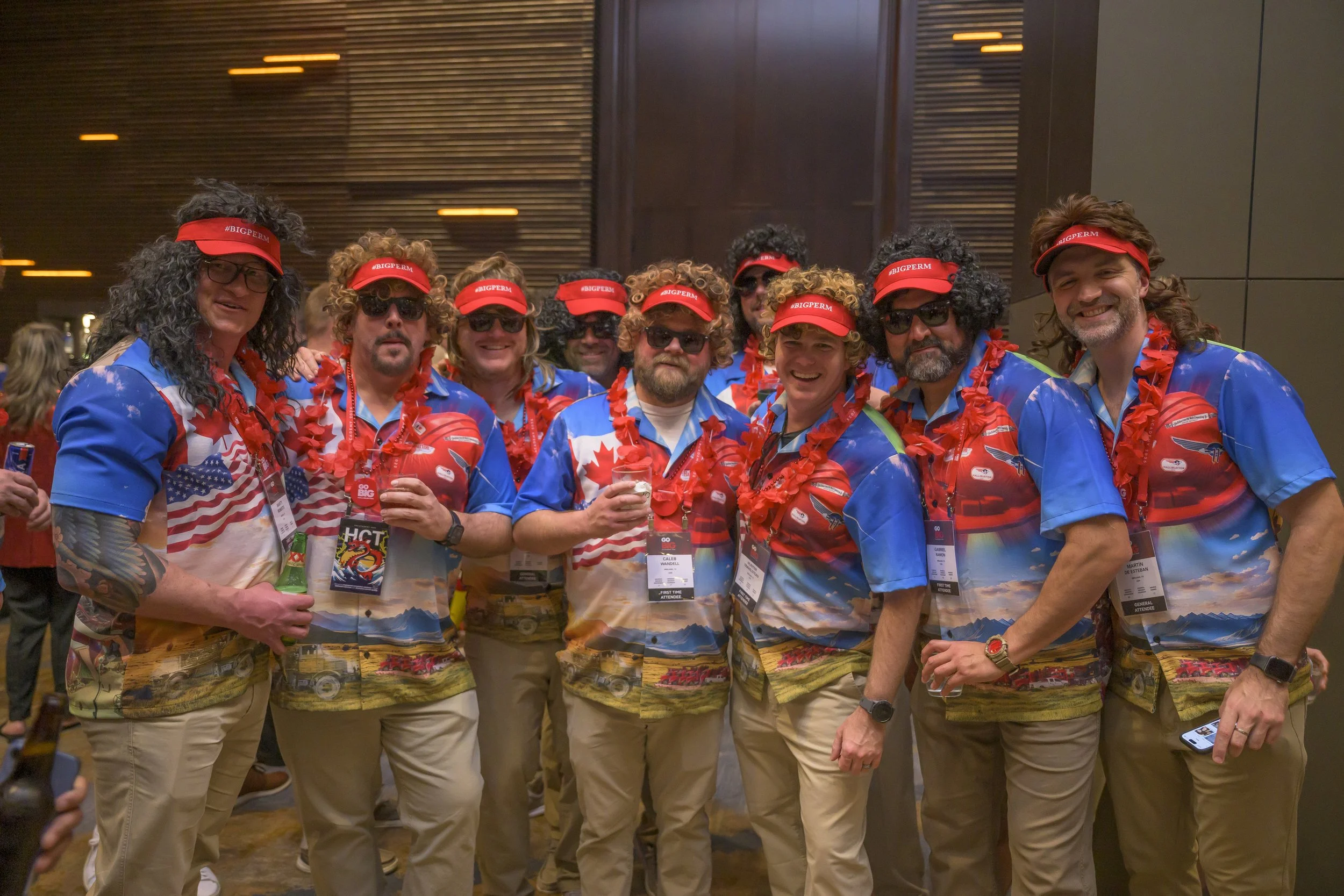 Group of men wearing matching blue and colorful patterned shirts, red visors, and leis at a conference or event.