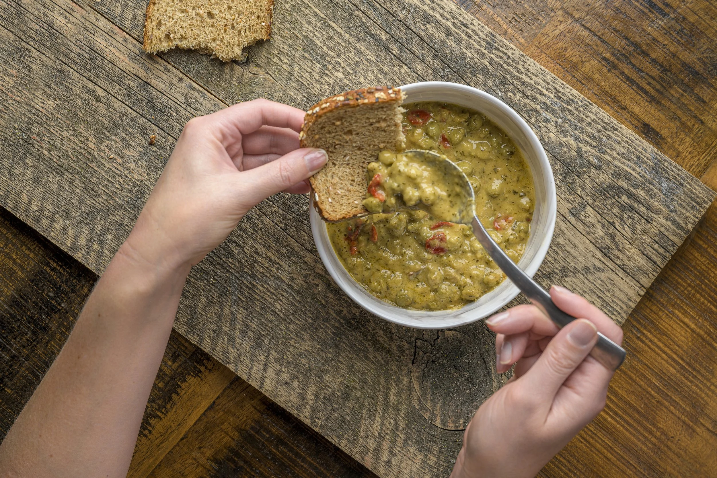 A person holding a slice of bread and dipping it into a bowl of greenish soup or sauce with chunks and red peppers on a rustic wooden table.