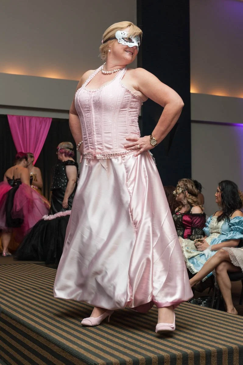 Woman in a pink ball gown and mask dancing on a runway during a dress-up event, with audience members in fancy dresses watching in the background.