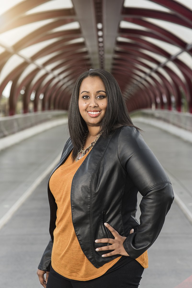 A woman with shoulder-length black hair wearing a black leather jacket over a mustard-colored top, standing on a bridge with an arched red metal framework in the background, smiling at the camera.
