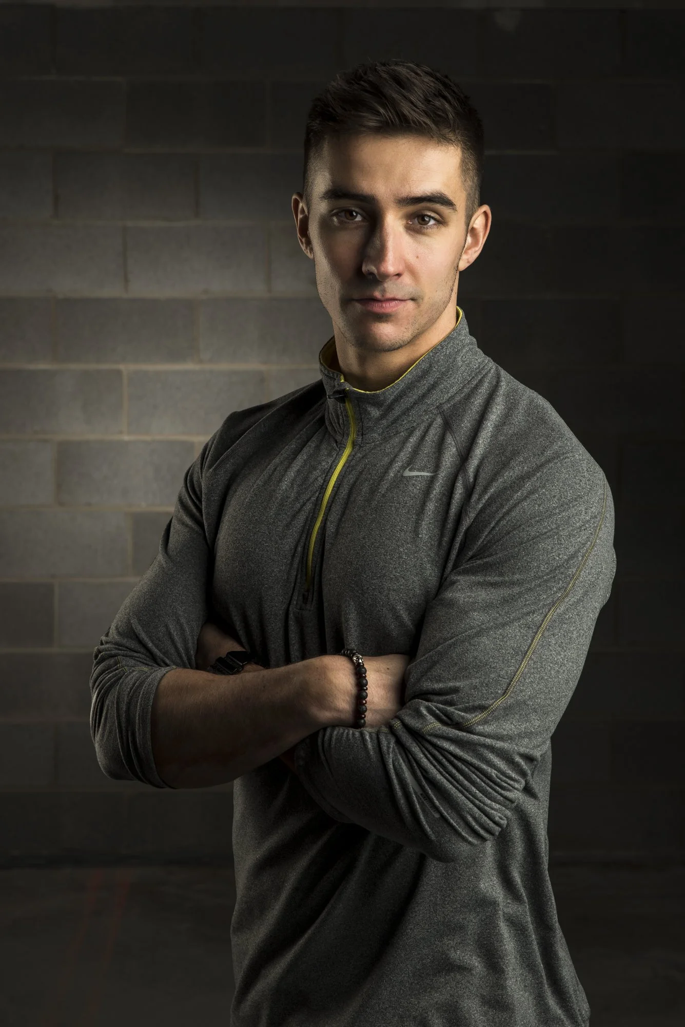 Young man with short brown hair wearing a dark gray athletic jacket with yellow accents, standing with arms crossed against a brick wall background.
