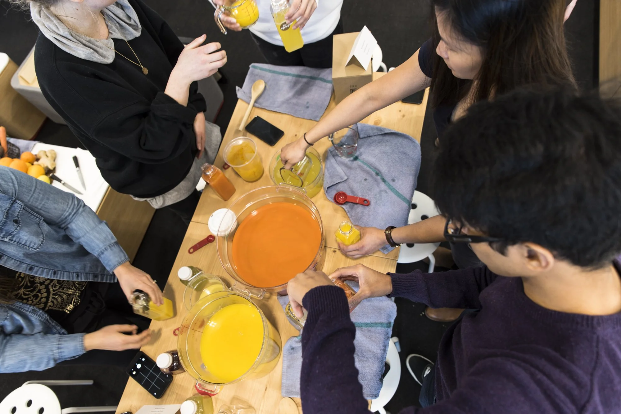 A group of people gathered around a wooden table, making fruit juices. The table has large pitchers of orange and yellow juice, small bottles of flavoring, and some glasses. The scene appears to be in a cozy indoor setting.