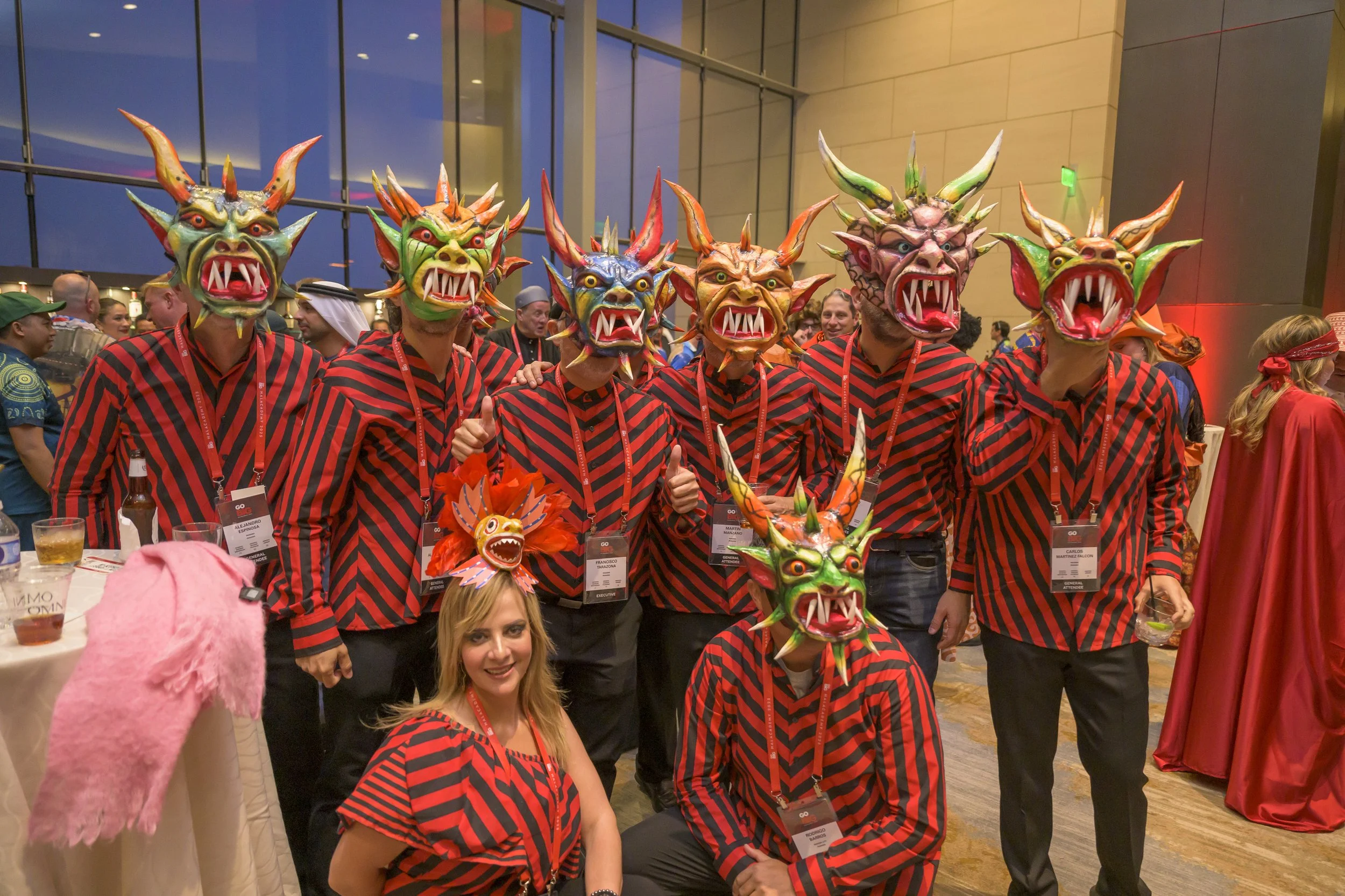 Group of people wearing colorful dragon masks and matching red and black striped shirts at a convention or event.