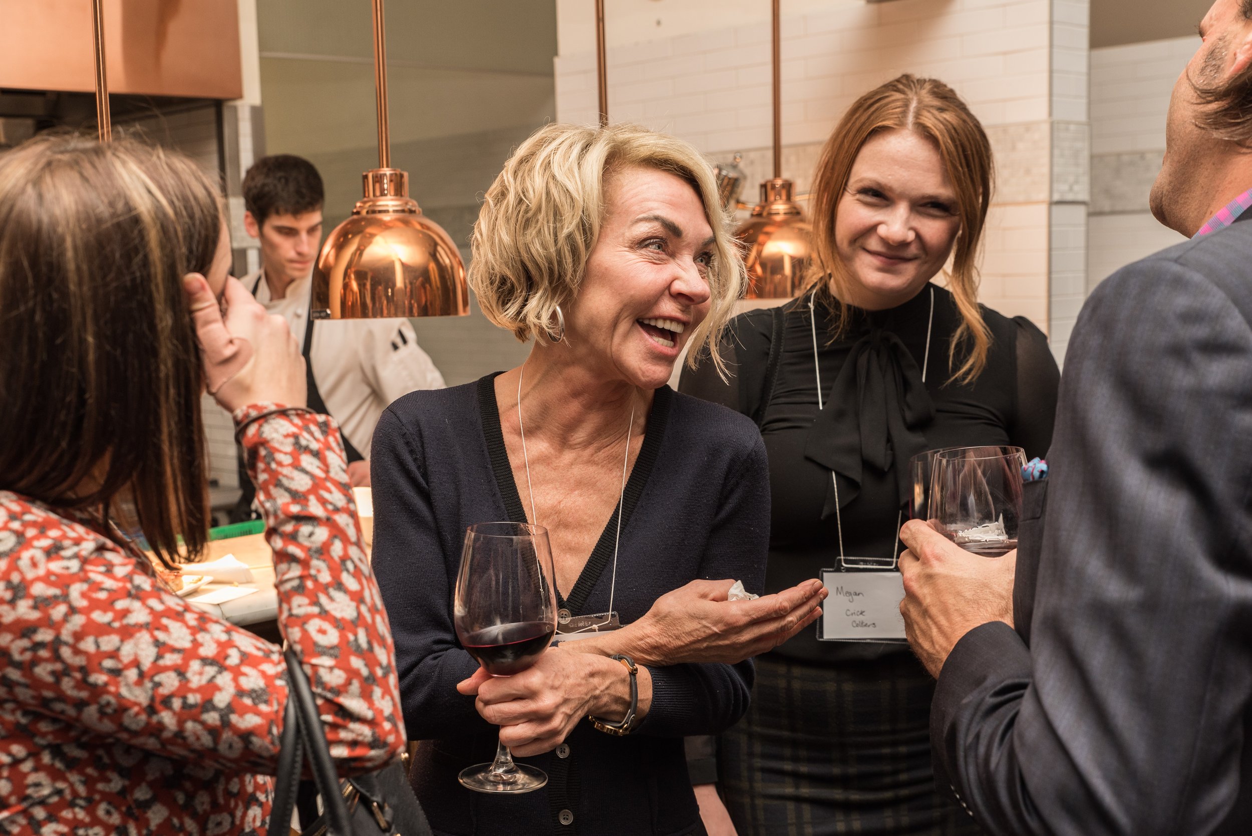 Group of people at a social event, engaging in conversation and holding glasses of wine, in a warmly lit indoor setting.