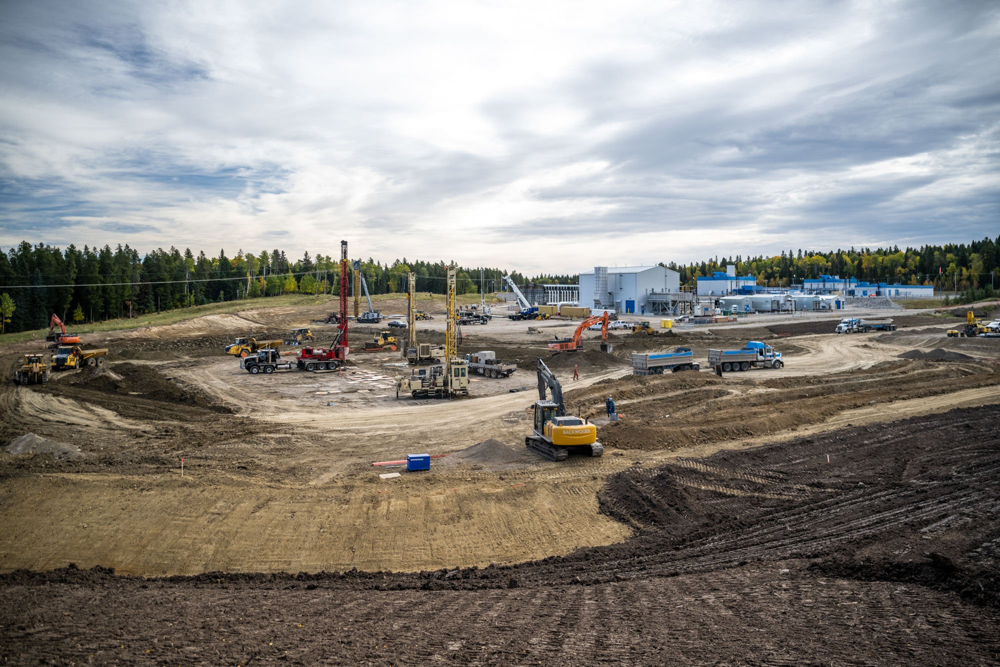 Construction site with heavy machinery, cranes, trucks, and workers, with a building in the background and a cloudy sky overhead.