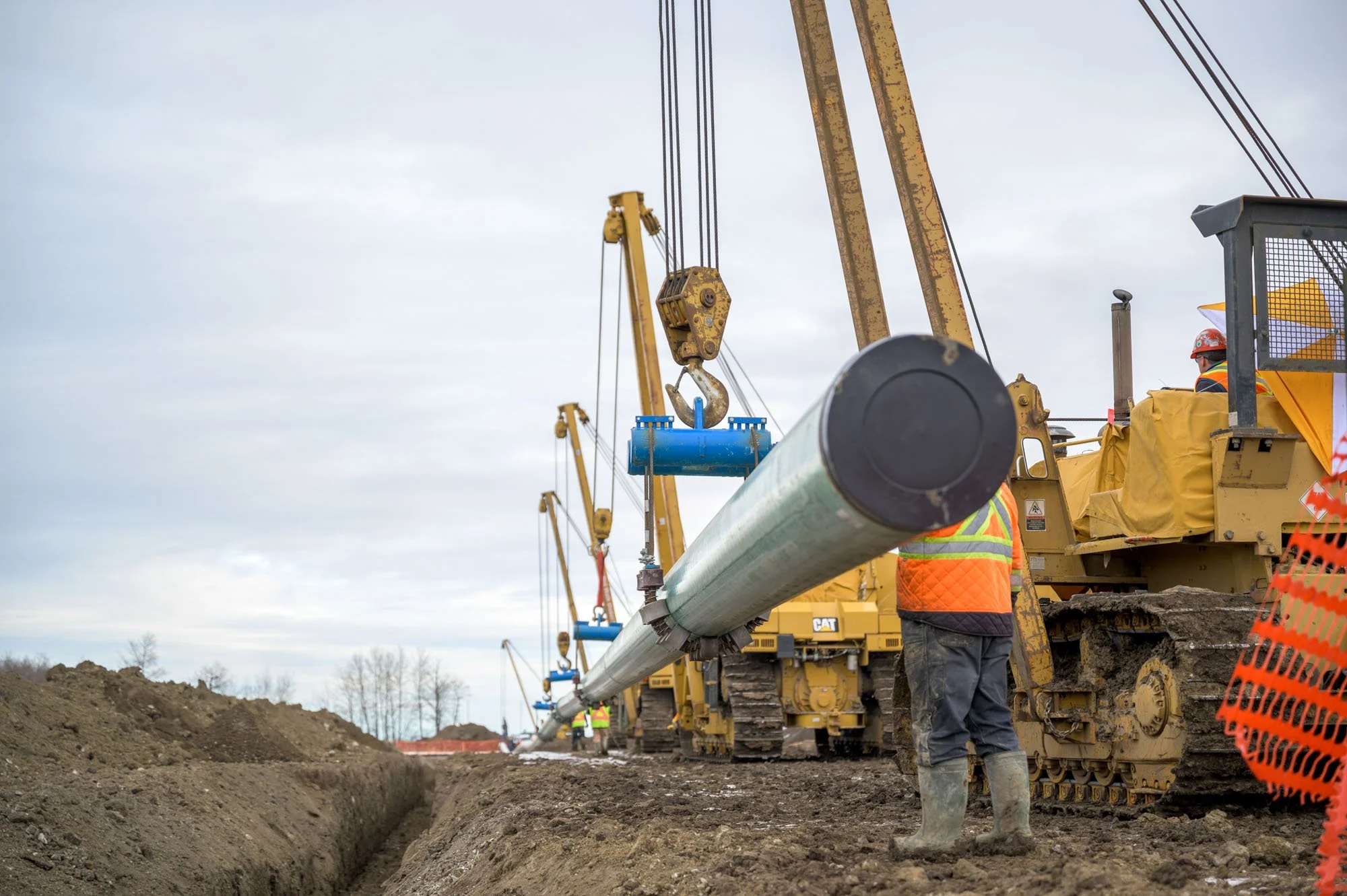 Construction workers operating heavy machinery on a construction site with large pipes being installed, and cloudy sky overhead.