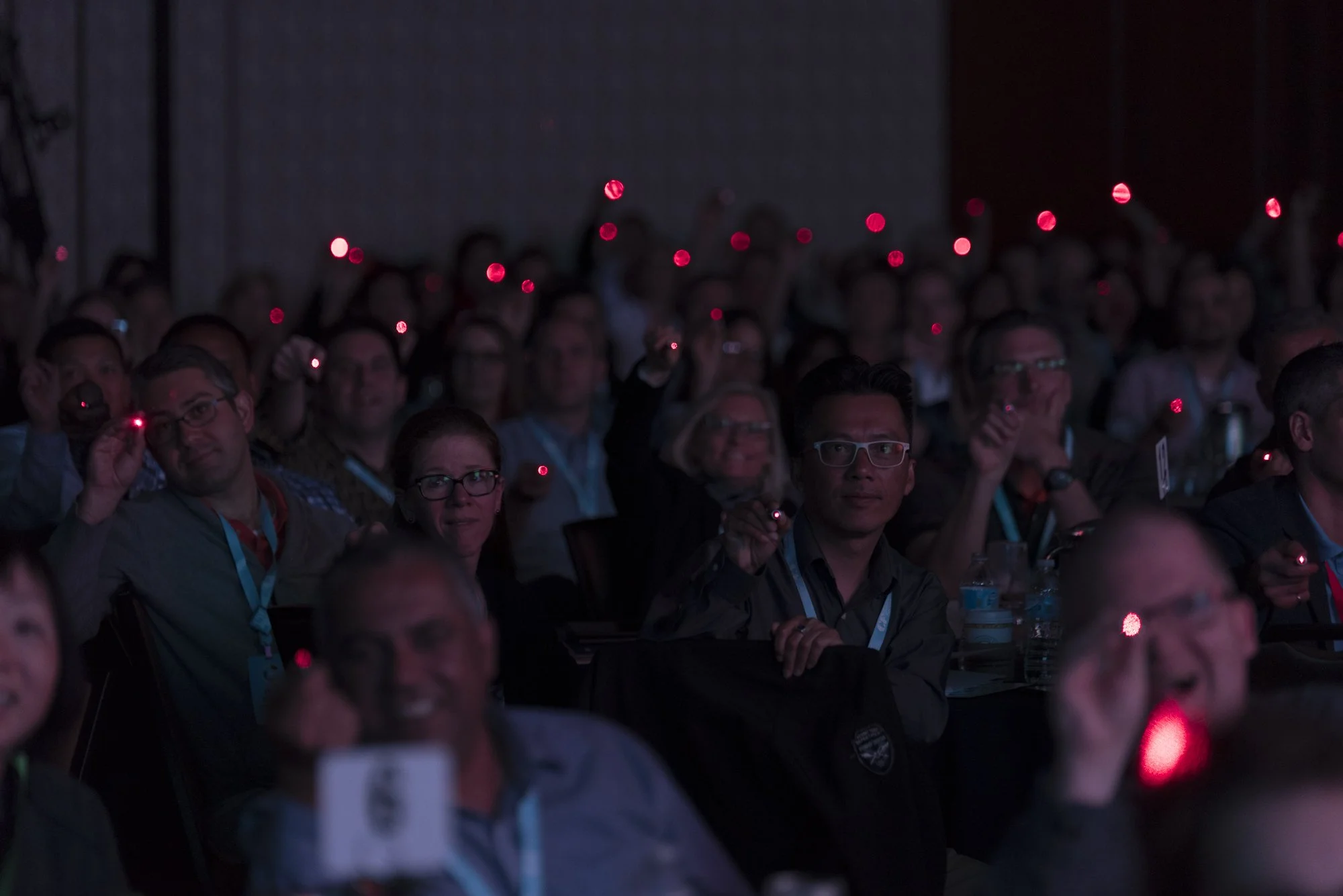 Audience at a conference holding red LED lights, sitting in a dark room, watching a presentation.