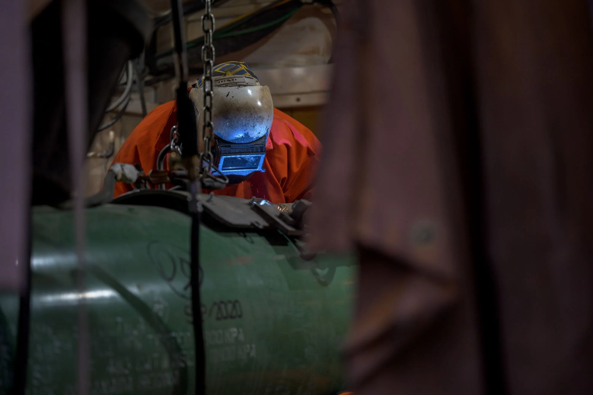 A welder wearing protective gear, including a helmet with a face shield and orange jacket, welding metal in an industrial workshop while surrounded by chains and equipment.
