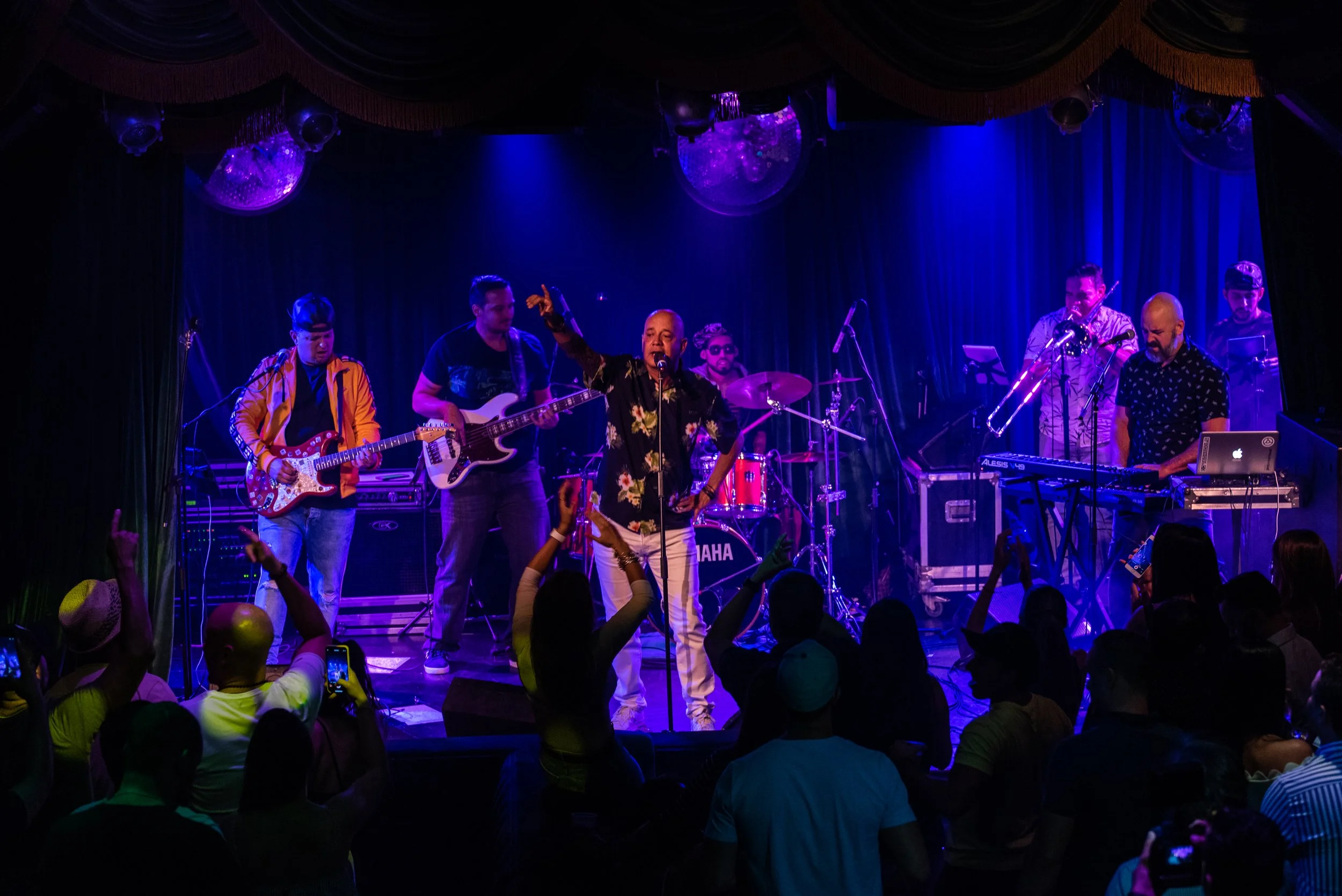 A live music performance on stage with six musicians playing instruments including guitar, bass, drums, keyboard, and trumpet, while singing and engaging with the audience in a dark venue with colorful stage lighting.