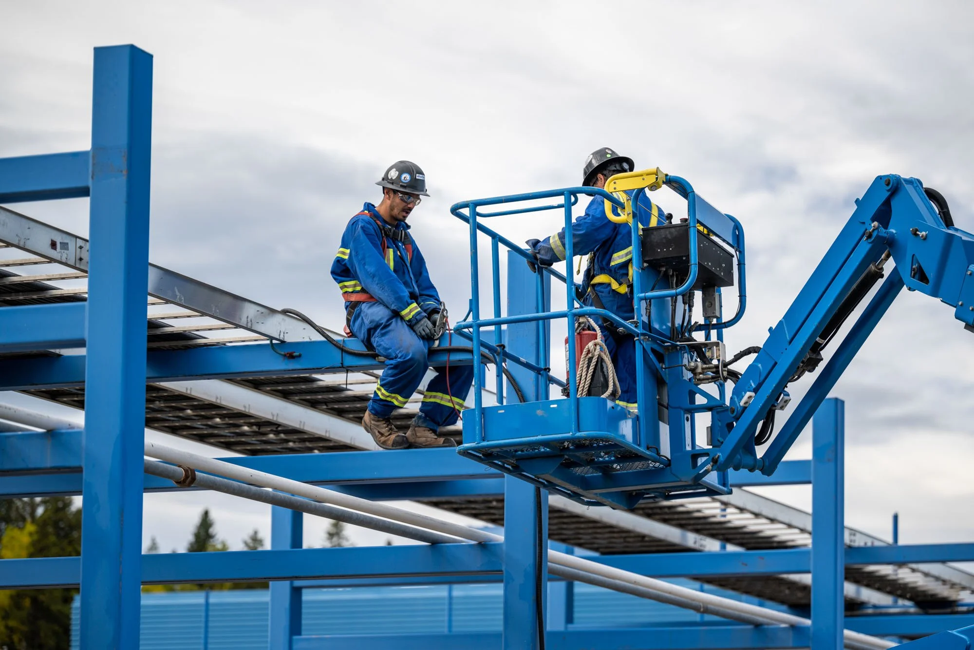 Two construction workers in blue safety gear and helmets working on a blue steel structure using a cherry picker lift.