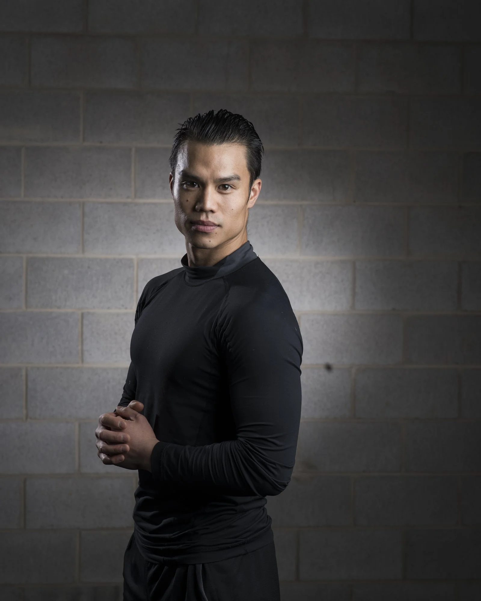 A young man in black athletic clothing standing against a gray brick wall, looking confidently at the camera with hands clasped in front of him.