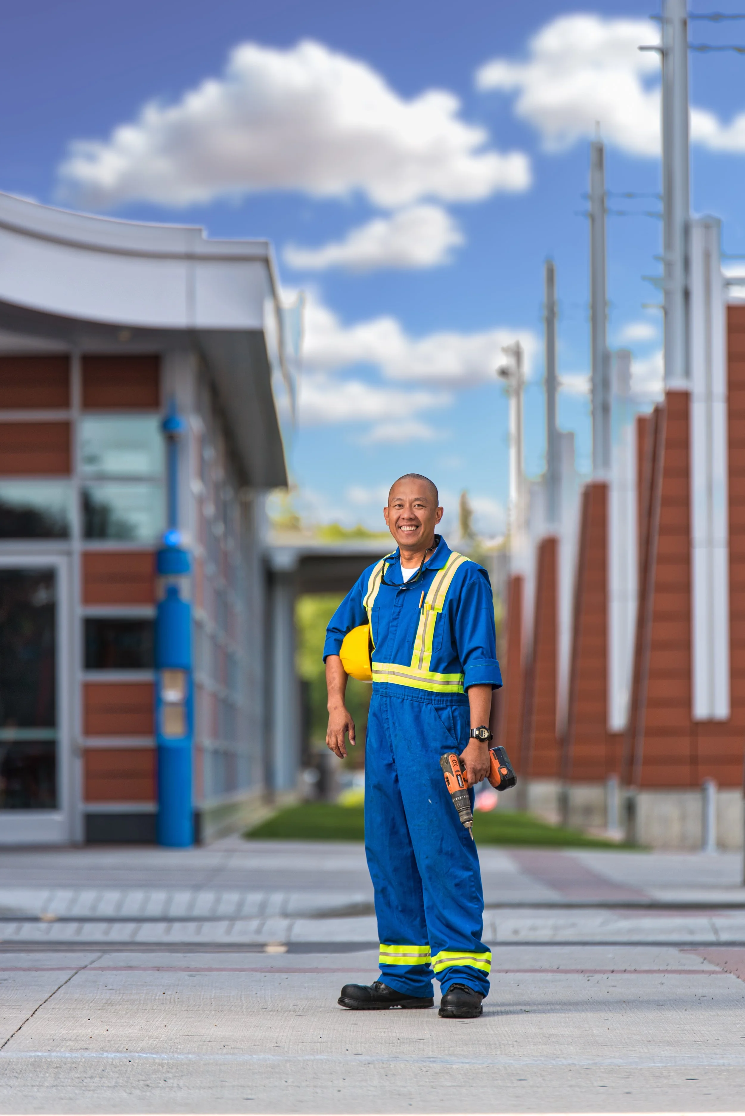 A smiling construction worker in a blue uniform standing on a sidewalk with a drill in hand and holding a yellow safety helmet, with buildings and a partly cloudy sky in the background.
