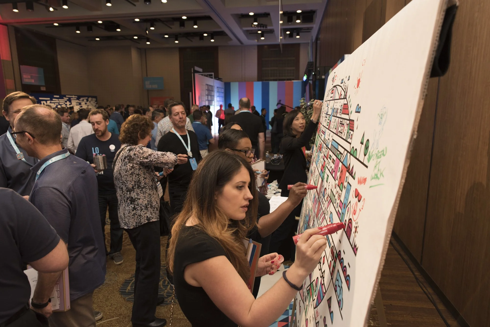 People participating in a collaborative drawing activity at a busy conference or event, with some individuals drawing on a large whiteboard filled with colorful illustrations, in a spacious room with a high ceiling and patterned walls.