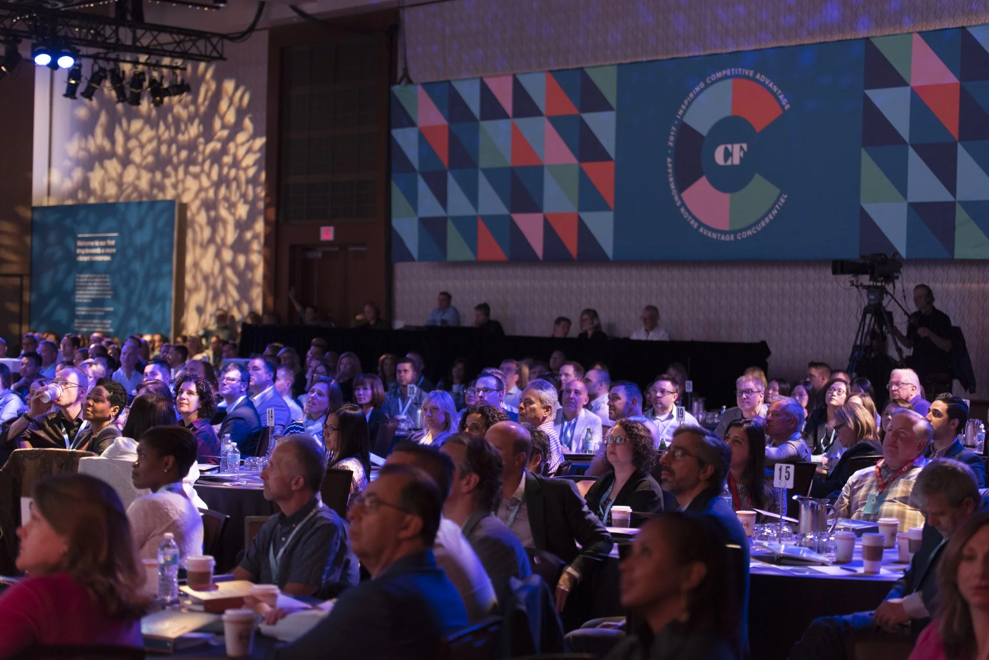 Crowd of business professionals attending a conference, sitting at tables with drinks and taking notes, in a large, dimly lit ballroom with colorful banners and a camera operator filming.