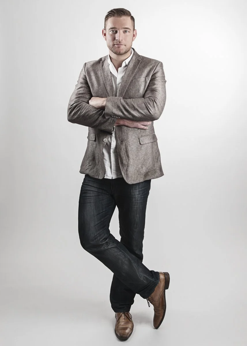 A man with short brown hair and a beard standing against a plain white background with arms crossed, wearing a gray blazer, white shirt, dark jeans, and brown dress shoes.